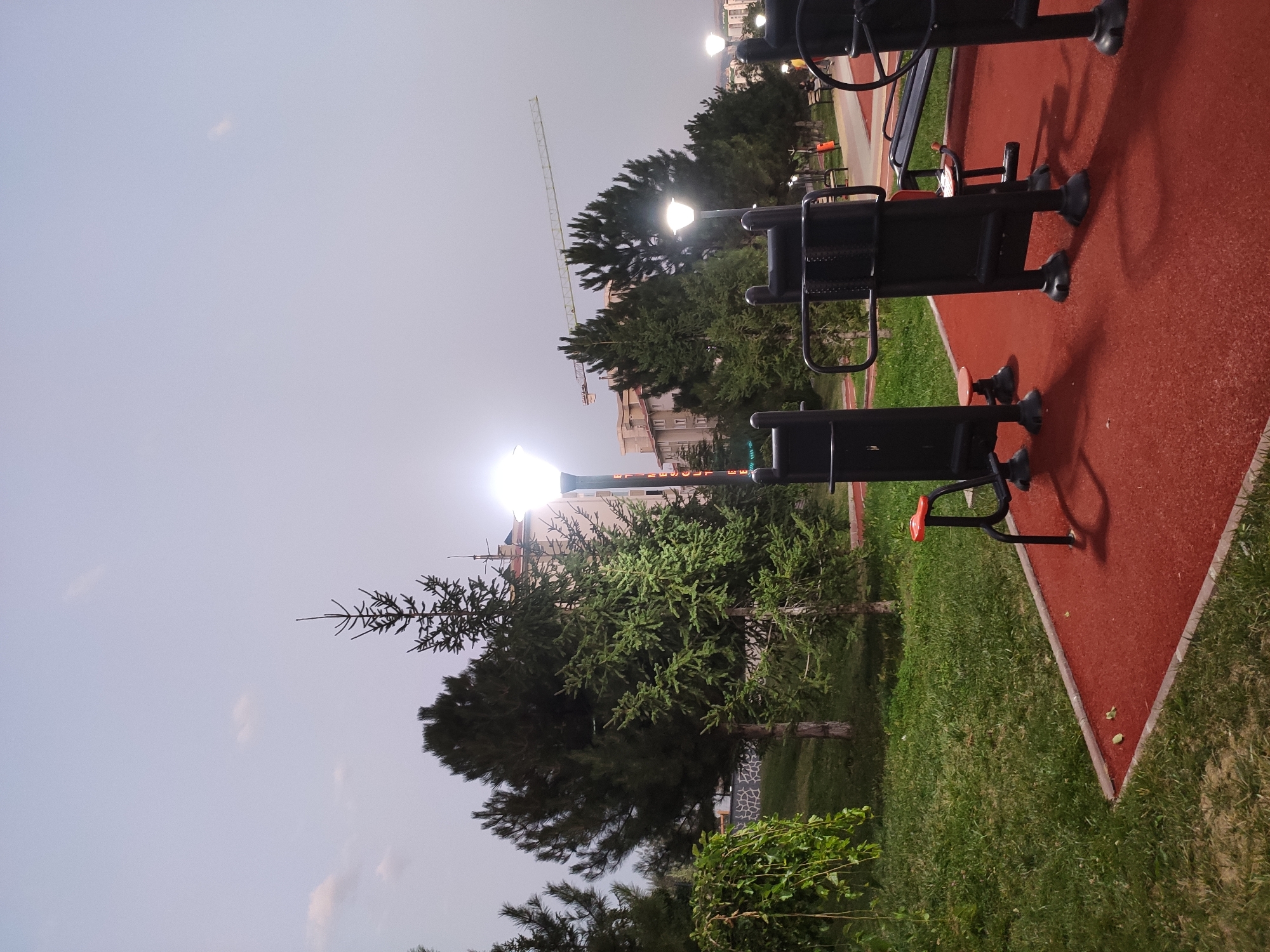 The image captures an outdoor fitness area in Etimesgut, Turkey, during twilight. In the foreground, red rubberized ground surfaces are visible, with several black outdoor exercise machines situated on them. These machines appear to be made of metal and include various components for strength training. To the left, a grassy area with trees and foliage is present, including a weeping willow-like tree. In the background, tall coniferous trees and residential buildings are visible. A construction crane is also present in the distance, suggesting ongoing development in the area. Streetlights illuminate the scene, casting shadows from the exercise equipment. The sky is a muted gray, indicating the transition from day to night. No people are clearly visible in the frame, though hints of activity might be perceived in the distance. The overall atmosphere is serene and functional, typical of a public recreational space.