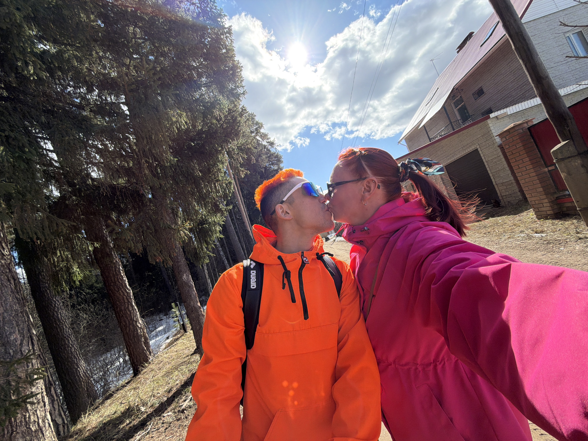 A photo depicts a couple kissing outdoors. The person on the left is wearing an orange jacket with a black backpack and has orange hair and white-framed sunglasses. The person on the right is wearing a pink jacket and glasses, with their hair pulled back in a ponytail. The photo appears to be taken on a sunny day, with tall trees on the left side and a building on the right. The location is Chaikovsky, Russia, based on metadata.