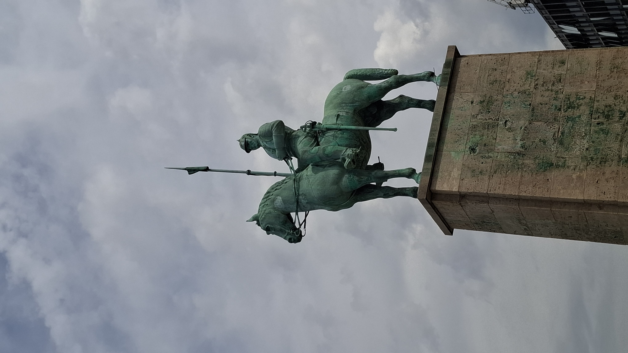 An equestrian statue, weathered to a greenish patina, depicts a figure in armor riding a horse. The rider holds a tall spear upright, its tip pointed towards a cloudy sky. The statue is mounted on a tall, rectangular stone pedestal, which appears textured and aged, with visible signs of weathering. In the background, a modern building with dark glass windows and horizontal accents is partially visible on the right. The overall scene is a low-angle shot looking up at the monument against a backdrop of a grey, overcast sky. This monument is the equestrian statue of Kaiser Wilhelm I, located in front of the Cologne Central Station (Hauptbahnhof) in Cologne, Germany.