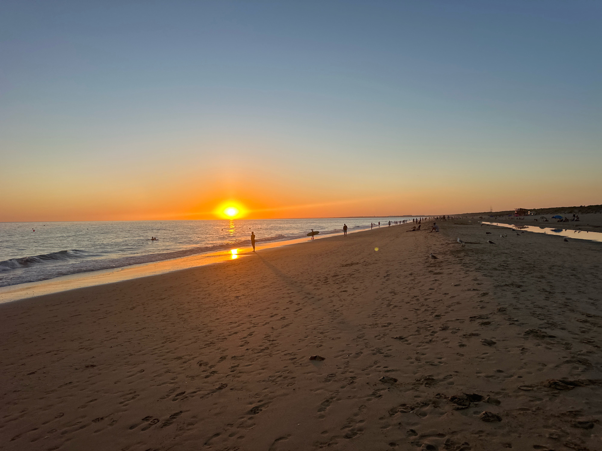 The image depicts a sunset scene at the beach in Punta Umbría, Spain. The sun is low on the horizon, casting a warm orange glow across the sky and reflecting on the ocean's surface. Waves gently lap the sandy shore, where numerous footprints are visible. Several people are scattered across the beach, some walking along the shoreline, others standing near the water, and a few appear to be swimming or wading in the sea. Further down the beach, a long line of individuals can be seen, suggesting a populated area. Birds, likely seagulls, are present on the sand. The sky transitions from a pale blue at the top to a gradient of yellow and orange near the horizon. The environment appears to be a coastal area with sand dunes visible in the distance.