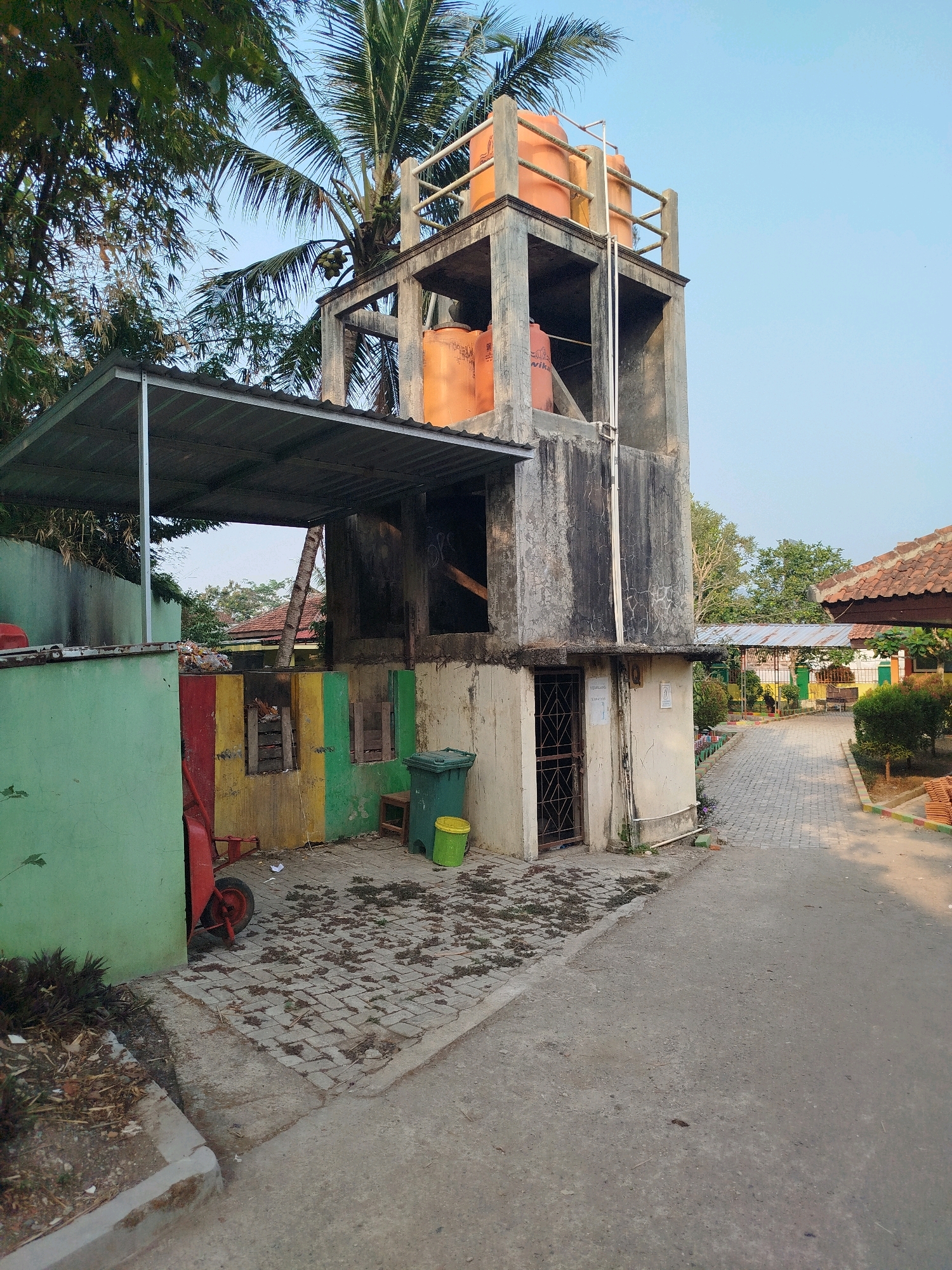 A concrete water tower stands beside a building with a green, red, yellow, and green painted wall. Orange water tanks are visible on top of the tower. A metal-roofed overhang extends from the building. In front of the building, a paved walkway leads away from the scene, bordered by greenery and another building with a tiled roof. A red wheelbarrow is parked next to the painted wall, and green and yellow trash bins are also present. The scene appears to be an outdoor area, possibly a school or community space, given the paved walkway and surrounding buildings. The sky is clear blue, and the lighting suggests it is daytime. The location is identified as Karangsambung, Indonesia.