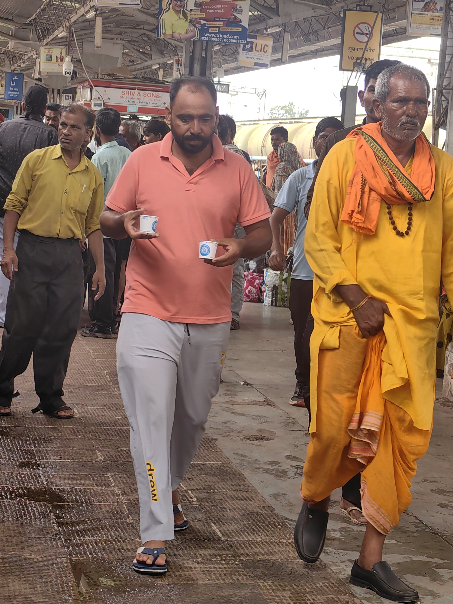The image depicts a busy scene at the Itarsi Railway Station in Itarsi, India. In the foreground, a man in a salmon-colored polo shirt and grey sweatpants is holding two small white cups. To his right, an older man in a bright yellow robe and an orange sash is walking, wearing a beaded necklace. Several other people are visible in the background, navigating the crowded platform. The environment includes overhead train station structures, signage, and luggage. The ground is wet, indicating recent rain or cleaning. The overall atmosphere suggests a typical bustling day at an Indian railway station.