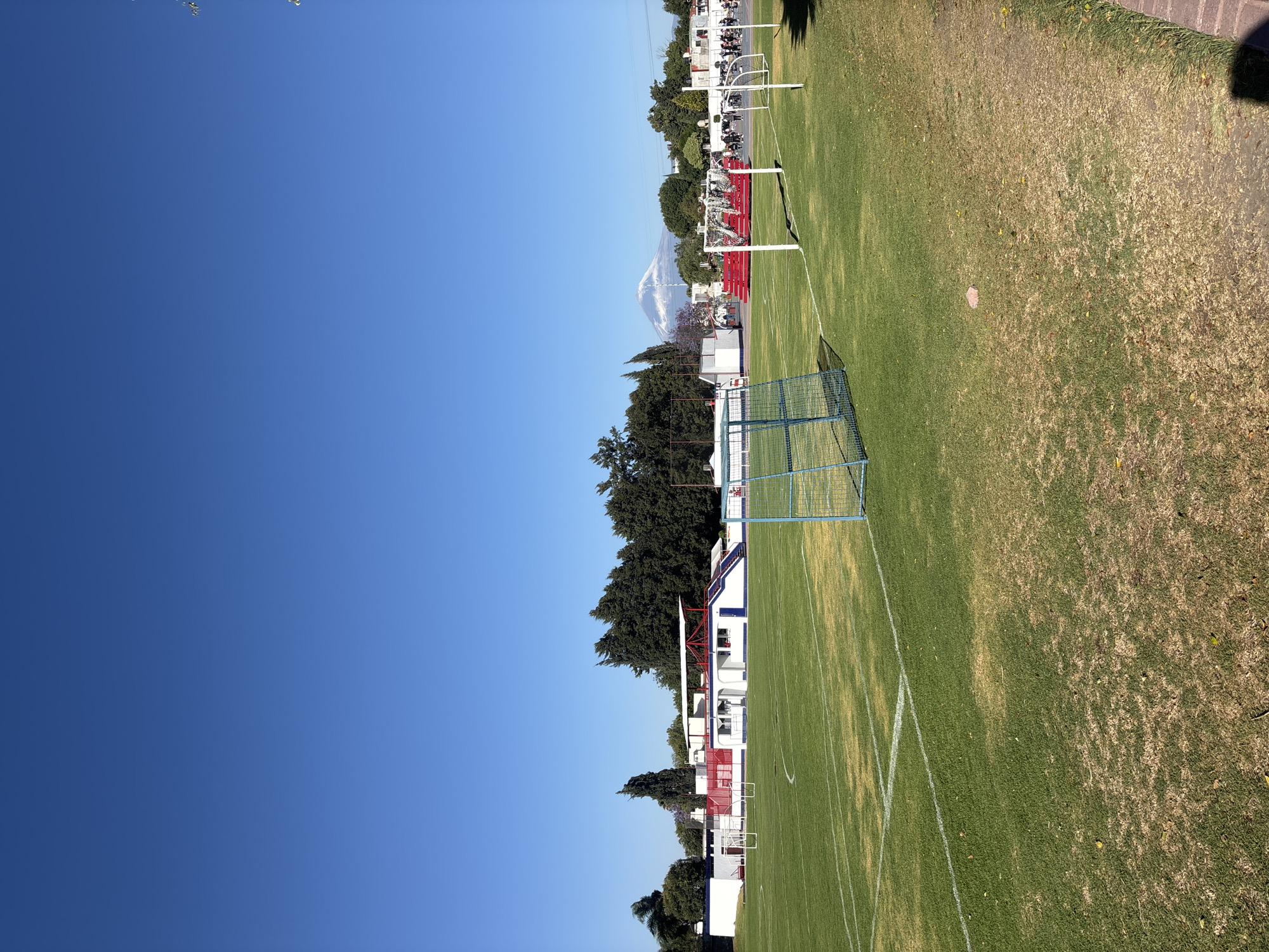 The scene captures an outdoor sports complex in San Bernardino Tlaxcalancingo, Mexico, under a clear, bright blue sky. The foreground features a grassy field, delineated by white lines consistent with a soccer pitch, exhibiting areas of both lush green and drier, brown vegetation. A light blue metal soccer goal with a net stands in the left foreground. Several white soccer goals are positioned further back on the field. In the midground, a white building with red roof accents and blue exterior stairs is visible on the left. To the right midground, red tiered seating is present. A group of approximately 10-15 individuals, predominantly in dark attire, is gathered near these red stands. The background is characterized by a dense line of trees, including conifers and one tree displaying purple blossoms. A prominent snow-capped mountain is visible above the tree line in the distant background. Sunlight illuminates the entire scene, indicating clear weather conditions.