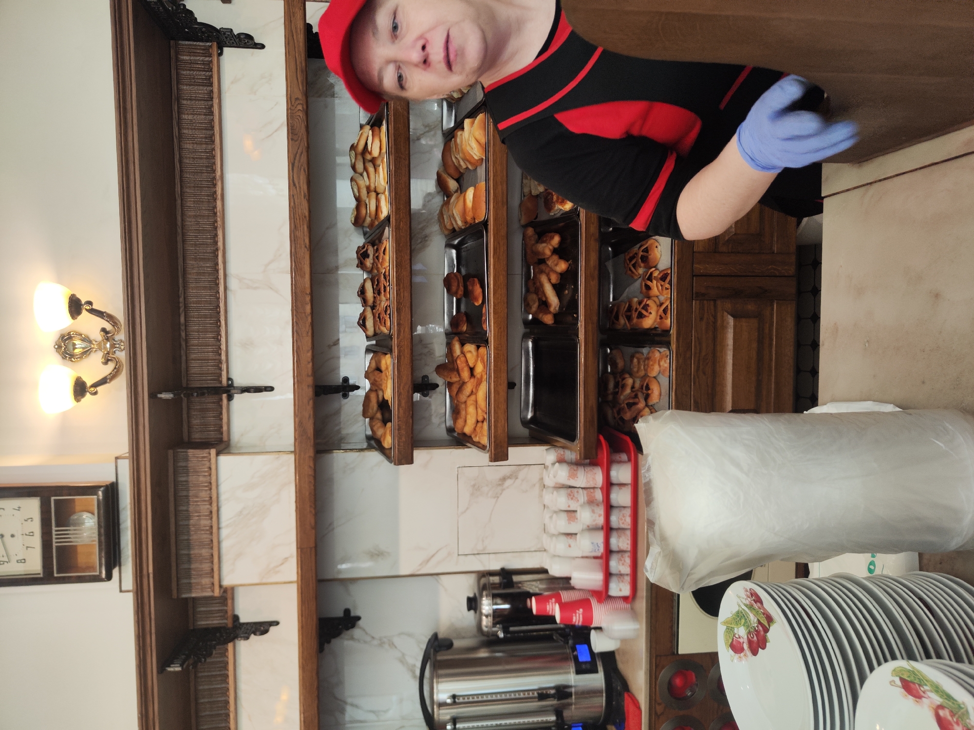 The photograph depicts a woman in a red cap and black and red shirt wearing blue gloves, standing in a bakery-like establishment. Shelves filled with baked goods are visible in the background, along with a lamp, a clock, a coffee machine, a cup holder, a plastic bag, and stacked plates. The location appears to be a shop, with a wooden cabinet and marble wall in the background.