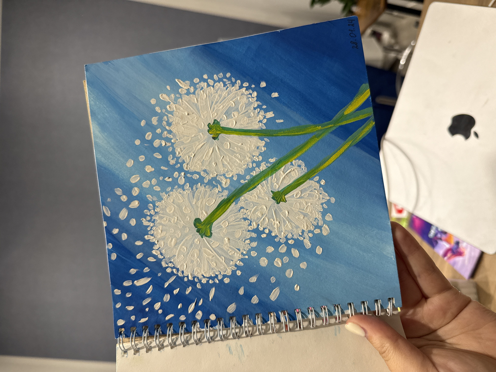 A person's hand, with a light-colored fingernail, holds an open spiral-bound sketchbook. The sketchbook displays a vibrant acrylic painting of three dandelion seed heads against a bright blue background with white flecks that suggest wind-blown seeds. The dandelions are depicted with textured white petals and green stems. In the bottom right corner of the painting, a date "28.01.24" is written in black ink. The background environment is blurred, showing a dark wall, a wooden table, and the corner of a white laptop with an Apple logo. There is no information in the image to suggest it is located in Murom, Russia.