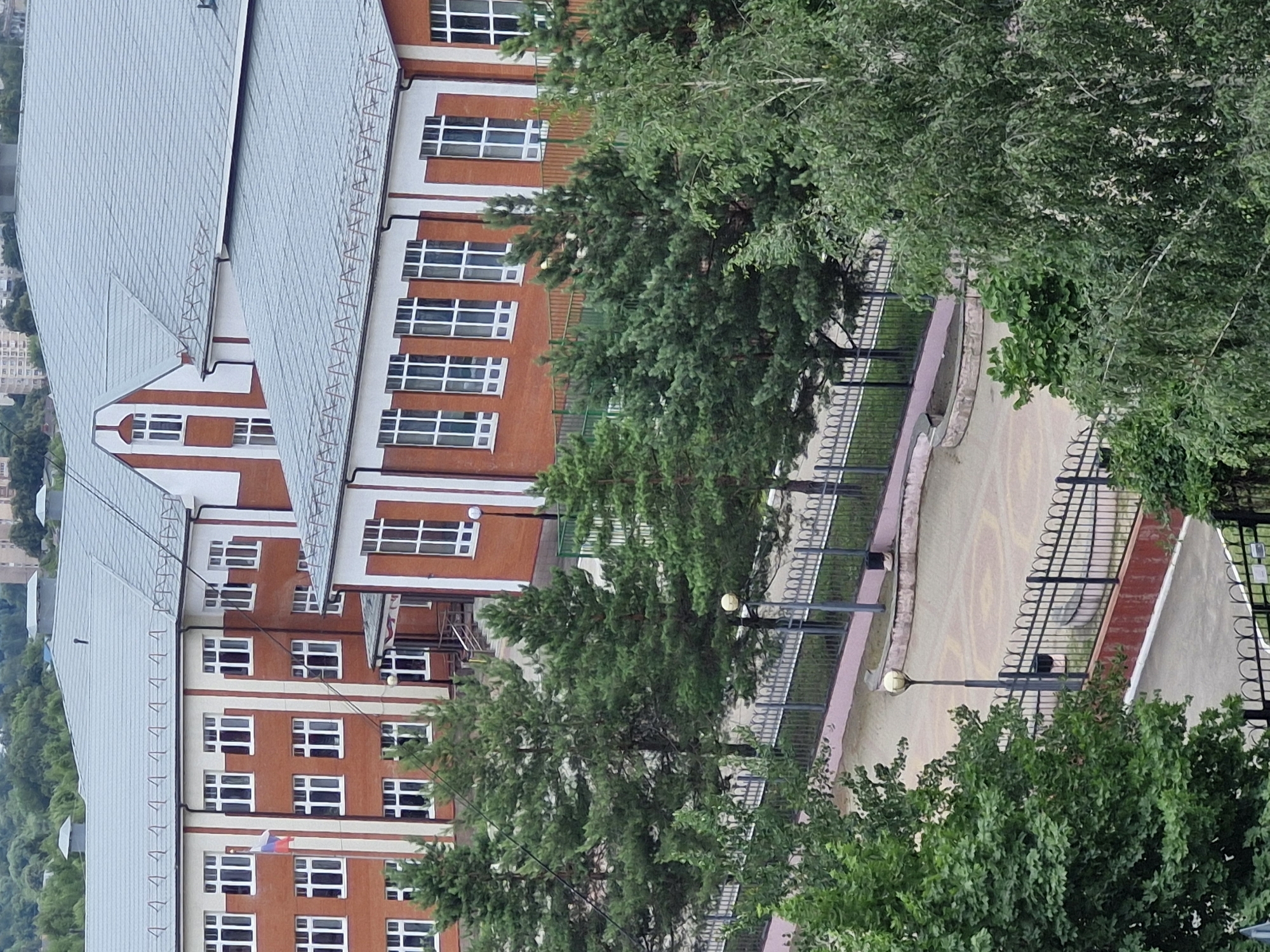 This is a medium-angle, vertical shot of a brick building with a light-colored roof, viewed from a slightly elevated perspective. The building appears to be a school or similar institution, characterized by its numerous windows with white frames. A Russian flag is visible on a pole on one of the building's floors.

The scene is dominated by lush green trees, which partially obscure the building and the surrounding grounds. A paved area, possibly a walkway or courtyard, is visible to the right, bordered by a decorative fence and a pinkish-red border. A lamppost with a golden fixture stands near the walkway. The overall setting suggests an outdoor environment during daylight hours, likely in a suburban or semi-rural area. There is no discernible text in the image. The weather appears to be overcast, with soft, diffused lighting. No people are visible.