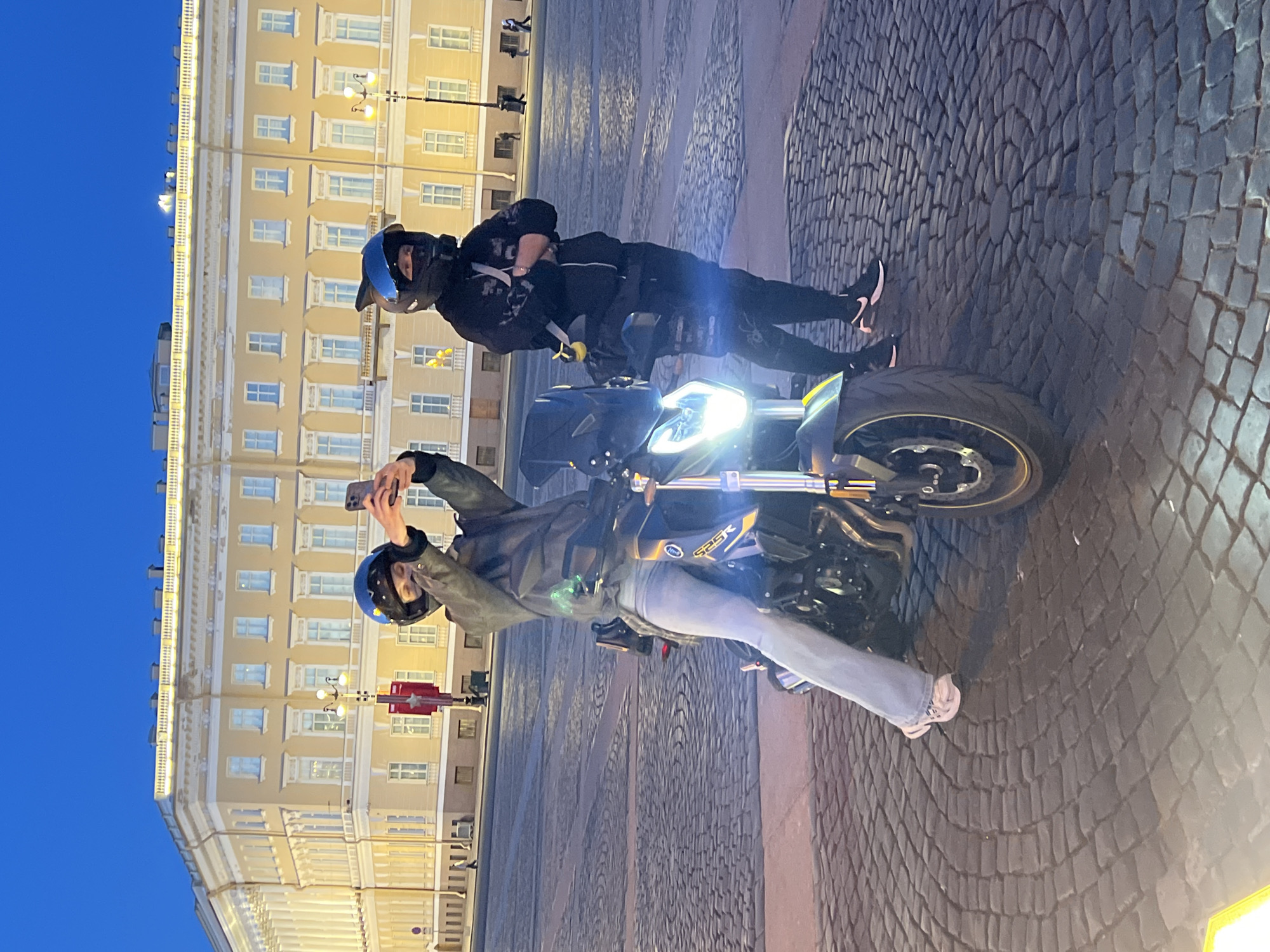 Two people are on a motorcycle, likely in Saint Petersburg, Russia. One person is on the motorcycle while taking a photo. The other person is standing next to the motorcycle. Both are wearing helmets and dark-colored attire. The motorcycle is black and green with a bright headlight. The scene is outdoors on a cobblestone surface, in front of a building with many windows and a yellow facade. The sky is visible and appears to be dusk or evening.