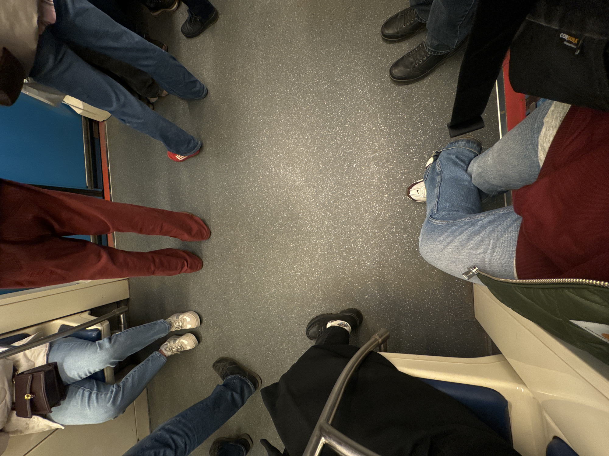 The image displays the interior of a Moscow Metro train carriage, viewed from a downward perspective, primarily focusing on the lower extremities of multiple individuals. The floor is covered in a speckled grey material. Visible train architecture includes portions of cream-colored plastic seat frames with blue upholstery, a silver-colored metal handrail, and a blue wall panel with a thin red stripe running horizontally near the floor.
Six distinct sets of legs and feet are visible, indicating multiple occupants:
1. **Top left:** A person seated, wearing light blue jeans, a white jacket, and a brown shoulder bag. Their feet are clad in white and beige sneakers.
2. **Top center:** A person standing, wearing dark red pants and matching dark red shoes.
3. **Top right:** A person standing, wearing dark blue jeans and red Adidas sneakers with white stripes.
4. **Mid-right:** Another person standing, wearing dark blue jeans and black lace-up shoes.
5. **Bottom left:** A person seated, wearing black pants, black chunky-soled shoes, and white socks with a dark pattern. Their left leg is partially obscured by a metal handrail.
6. **Bottom center/right:** A person seated, wearing light blue jeans, a dark red sweatshirt under a dark green quilted jacket, and white and maroon sneakers. Their left leg is crossed over their right.
The scene depicts a typical crowded public transport environment with people seated and standing in close proximity, but without direct interaction visible.