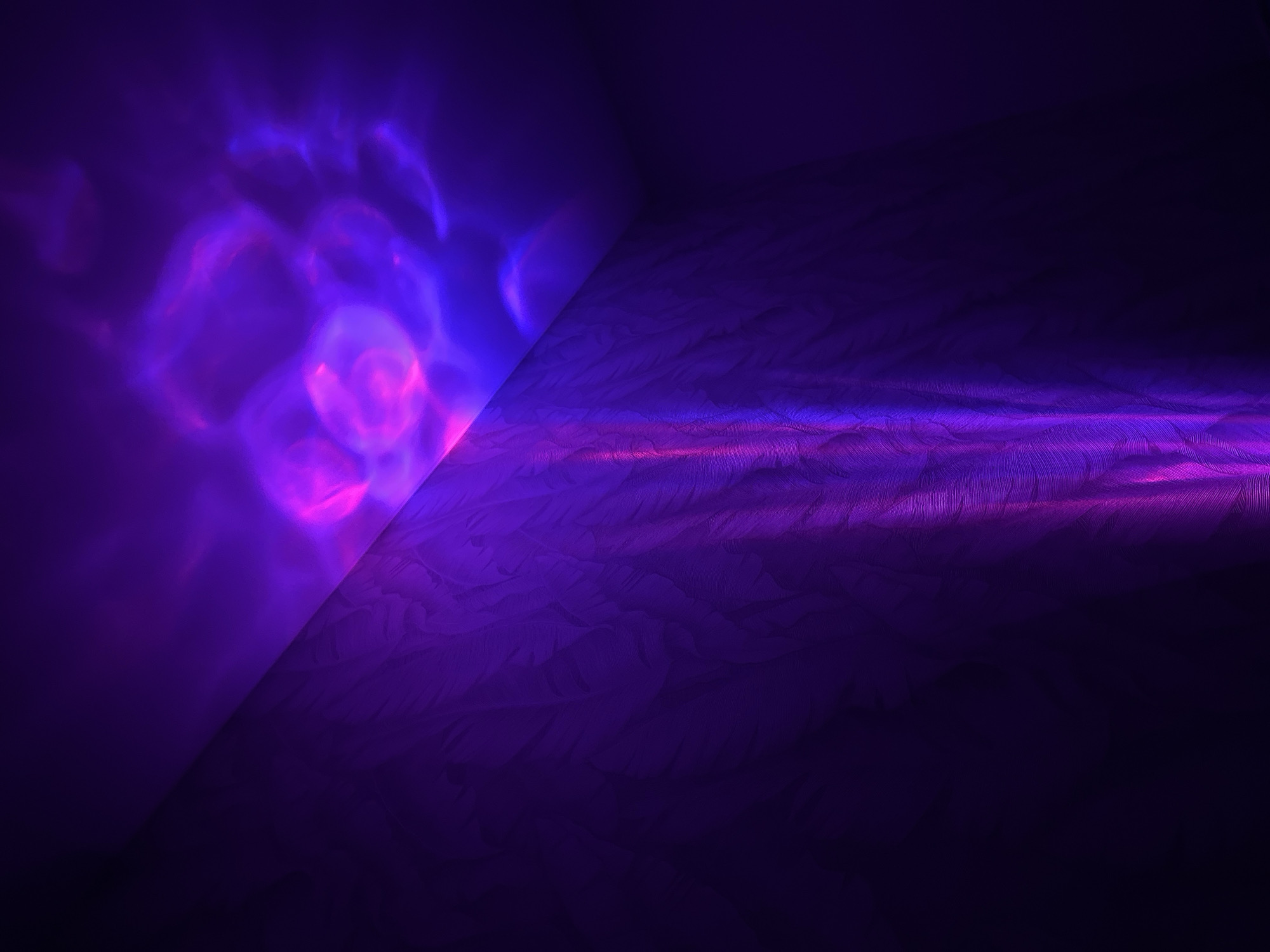 The image shows a corner of a room with a dark, textured purple wall on the right side and a smooth, lighter purple wall on the left. The left wall features blurry, bubble-like patterns of pink and blue light. A straight line diagonally separates the two wall textures. A horizontal band of pink light streaks across the right wall near the bottom. The overall lighting is dim and atmospheric. No people or specific objects are visible. There is no information to indicate the image was taken in Прикамский, Russia.