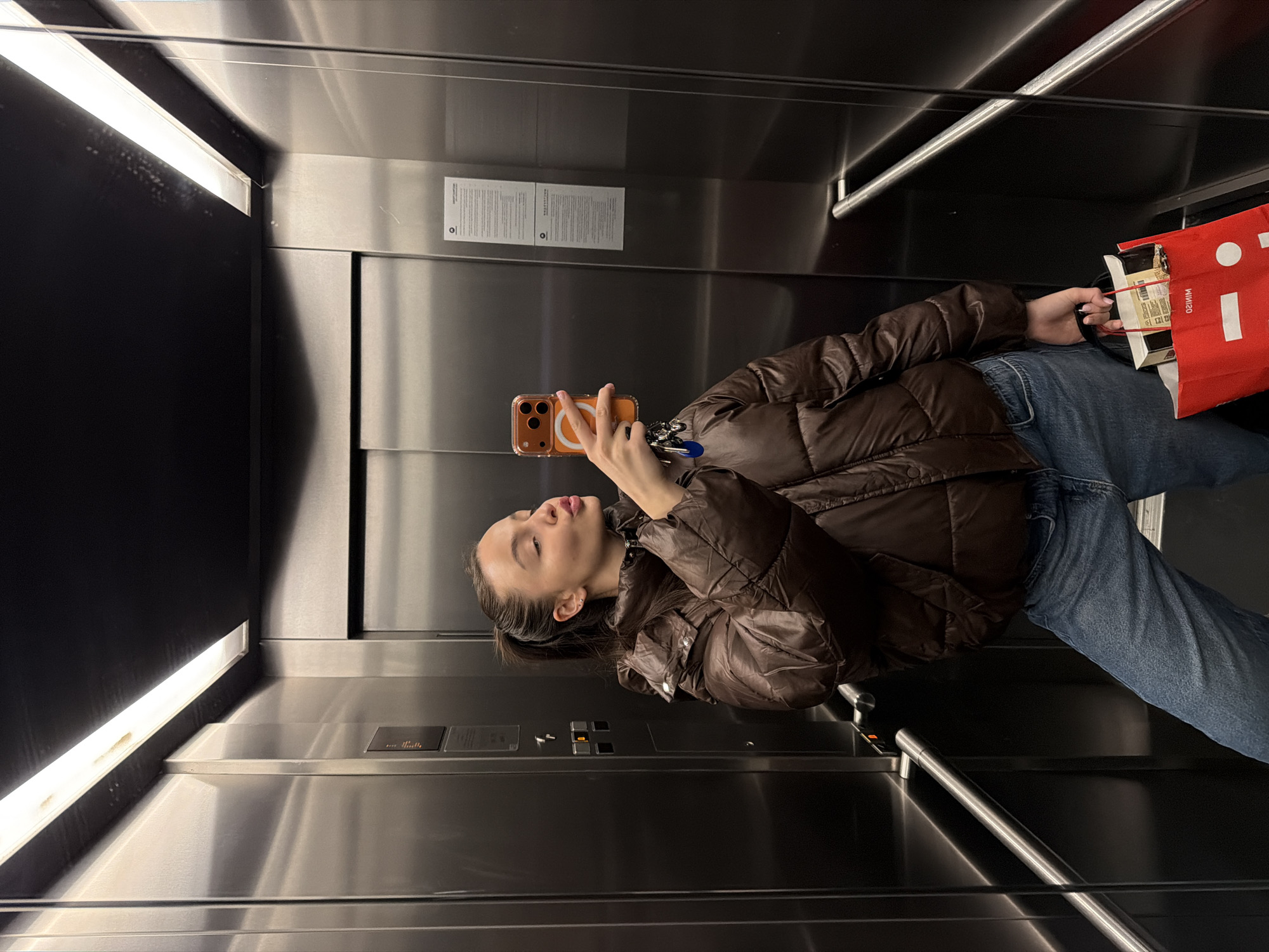 The image depicts a person taking a selfie in an elevator. The elevator has stainless steel walls with a dark ceiling and lighting. The person is wearing a brown puffer jacket and jeans, holding a phone in one hand and a red bag with groceries in the other. Based on the context of being taken in Sofia, Bulgaria, the elevator is likely in a residential or commercial building in Sofia.