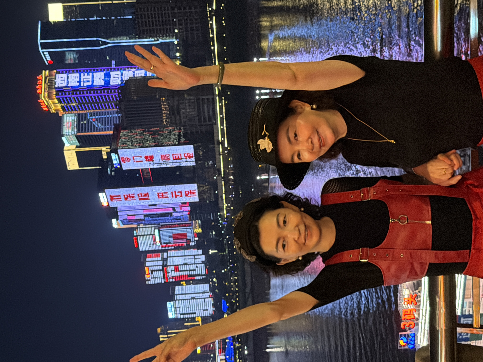 Two women pose for a photo at night in Yuzhong District, China, with the illuminated cityscape of Chongqing as a backdrop. The woman on the left wears a black top with a red leather harness and a black patterned headband. She is raising her left arm and waving. The woman on the right wears a black top, a black hat adorned with a flower, and pearl earrings. She is also raising her right arm and waving. Both women are smiling. The background features towering buildings with vibrant neon lights and reflections on the water. A railing is visible in the foreground.