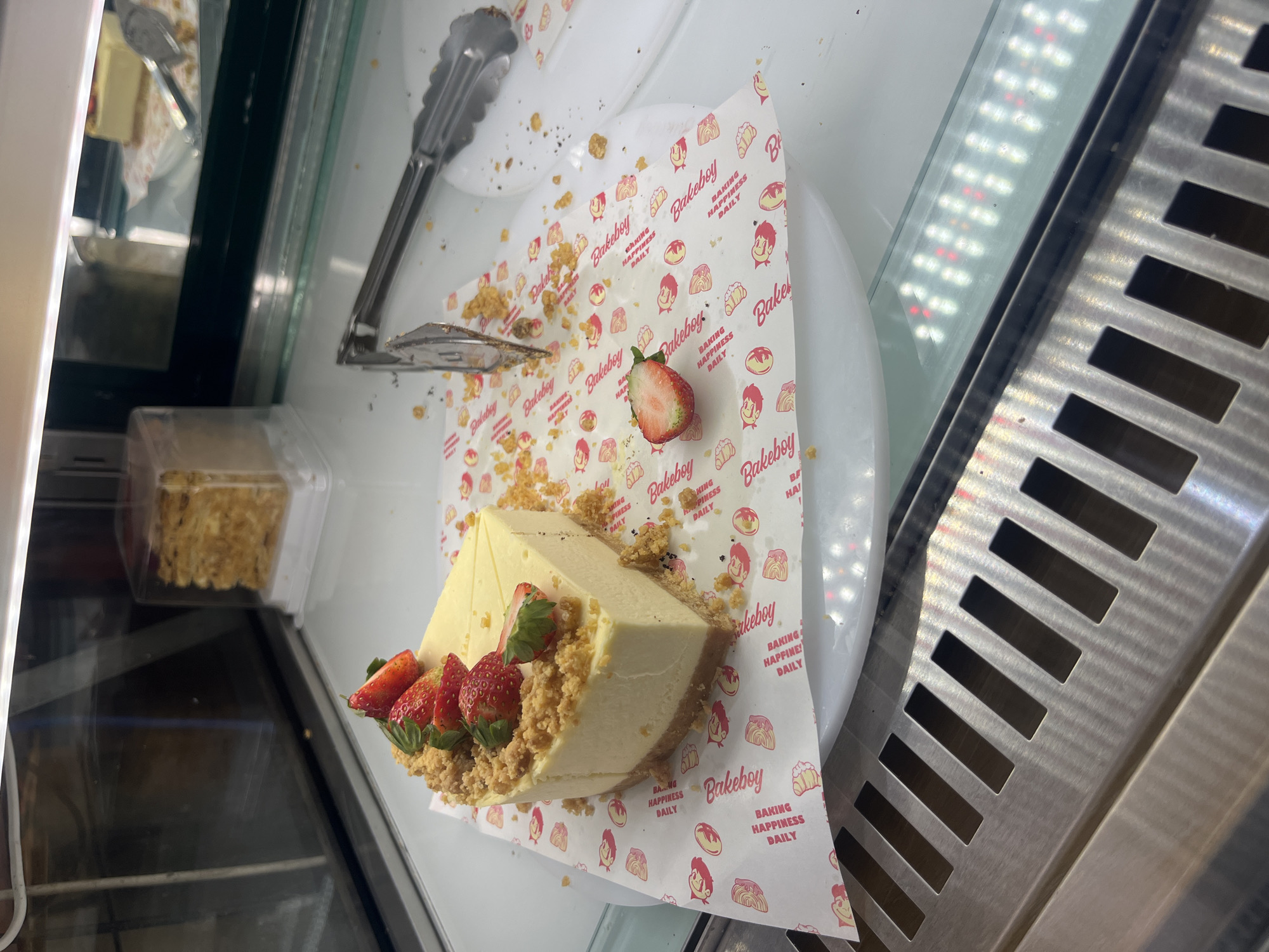 The image shows a slice of strawberry cheesecake displayed in a refrigerated display case, likely at a bakery or cafe. The cheesecake is garnished with fresh strawberries and sits on a decorative paper liner with the "Bakeboy" logo. Crumbs are scattered around the cake. A pair of metal tongs rests near the cake, presumably used for serving. The display case has a stainless steel exterior with ventilation slots. Another container of food is visible through the glass on the left side of the image. The location appears to be indoors.