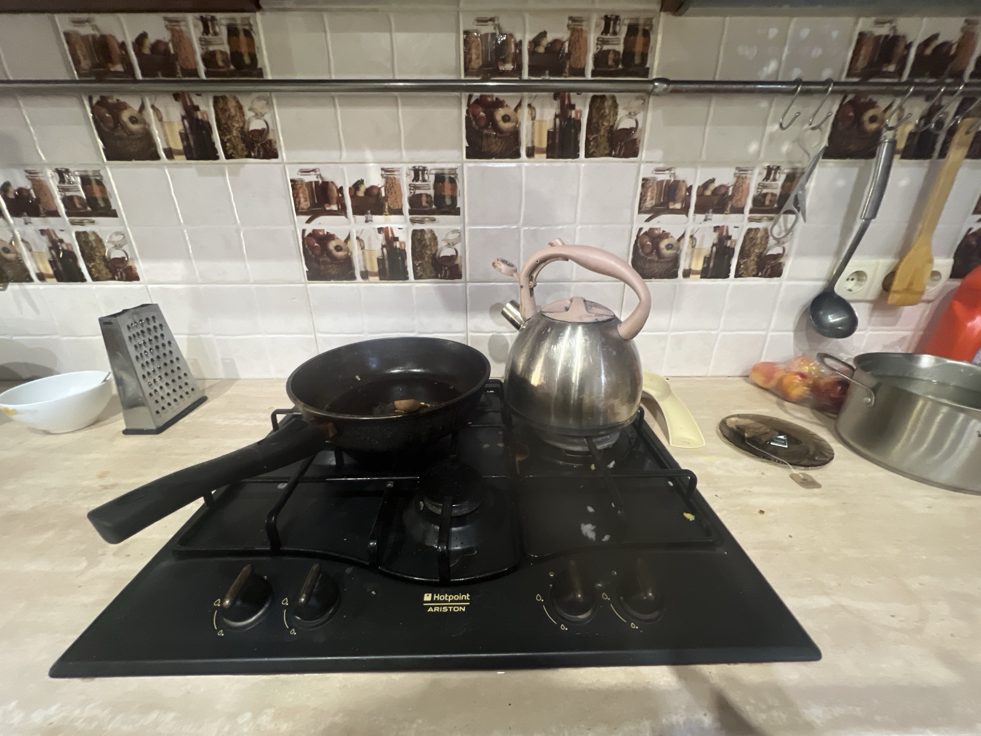 The image shows a kitchen countertop with a gas stovetop, a frying pan, and a kettle. The stovetop is black and has two knobs. The frying pan is black and contains some food. The kettle is made of metal and has a pink handle. A grater and a white bowl are also visible on the countertop. In the background, there is a tiled backsplash with a pattern of jars and other kitchen items. The kitchen appears to be in use, with cooking equipment laid out on the stovetop. There is no visible text in the image. The setting appears to be a domestic kitchen.