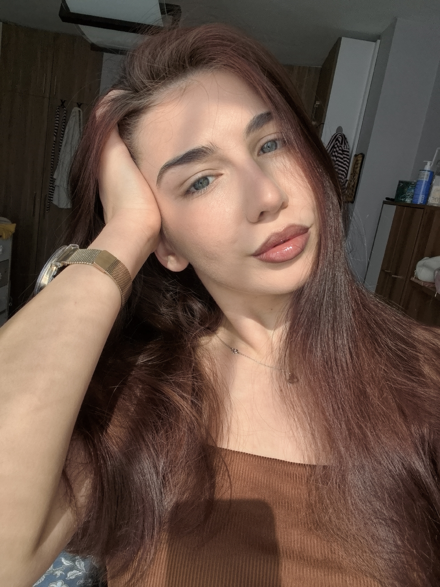 A young woman is captured in a close-up, chest-up portrait indoors, likely in a room in Stara Zagora, Bulgaria. She has long, dark reddish-brown hair that falls over her shoulders and striking blue-grey eyes. Her expression is calm and direct as she looks towards the camera.
She is wearing a brown ribbed top, possibly strapless or off-the-shoulder. On her left wrist, she sports a gold-toned mesh watch, and a delicate silver-toned necklace with a small pendant is visible around her neck. Her left hand is raised, with her fingers resting against her temple or cheek, giving a relaxed and somewhat pensive pose.
The scene is illuminated by strong natural sunlight, which casts distinct highlights and shadows across her face, hair, and arm, suggesting it is either morning or late afternoon. The background elements are somewhat out of focus but reveal an indoor setting with wooden furniture, possibly a closet or wardrobe, a light fixture on the ceiling, and on a shelf to the right, several bottles and a striped towel hanging from a hook. There is no visible text in the image.