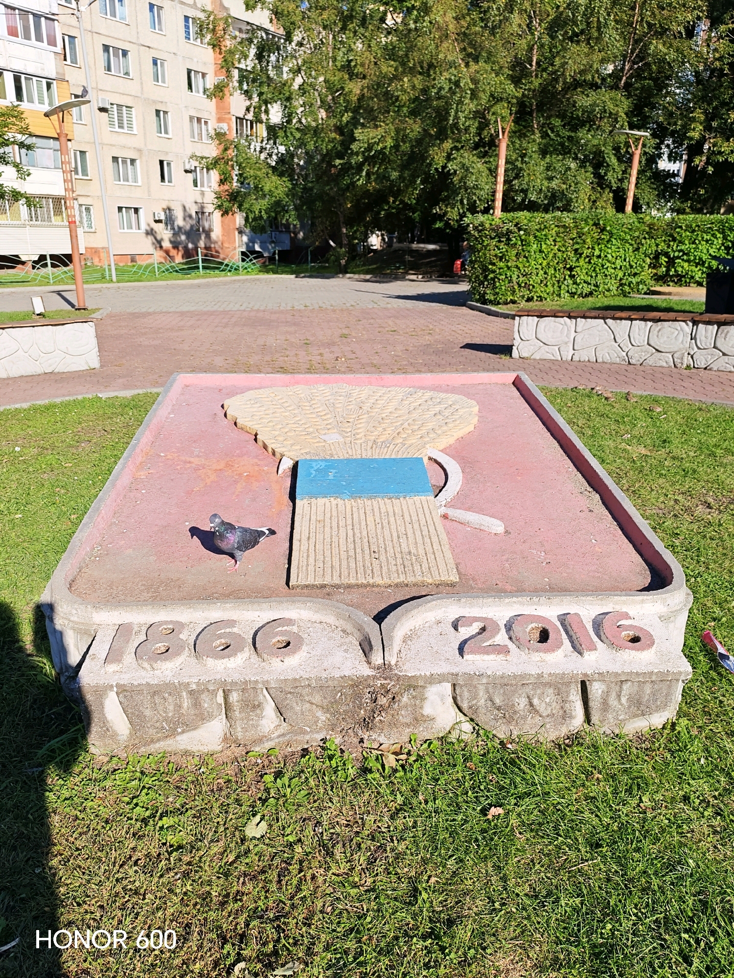 The image shows a concrete monument in a park in Ussurijsk, Russia. The monument is rectangular with rounded corners and features a raised relief depicting a bundle of wheat with a blue ribbon and a sickle. The numbers "1866" are carved on the left side of the monument and "2016" on the right side. A pigeon is perched on the monument. The surrounding area is a grassy lawn with a paved walkway in the background. In the background, there is a multi-story apartment building and trees. The lighting suggests it is daytime. The overall scene appears to be in good condition.
