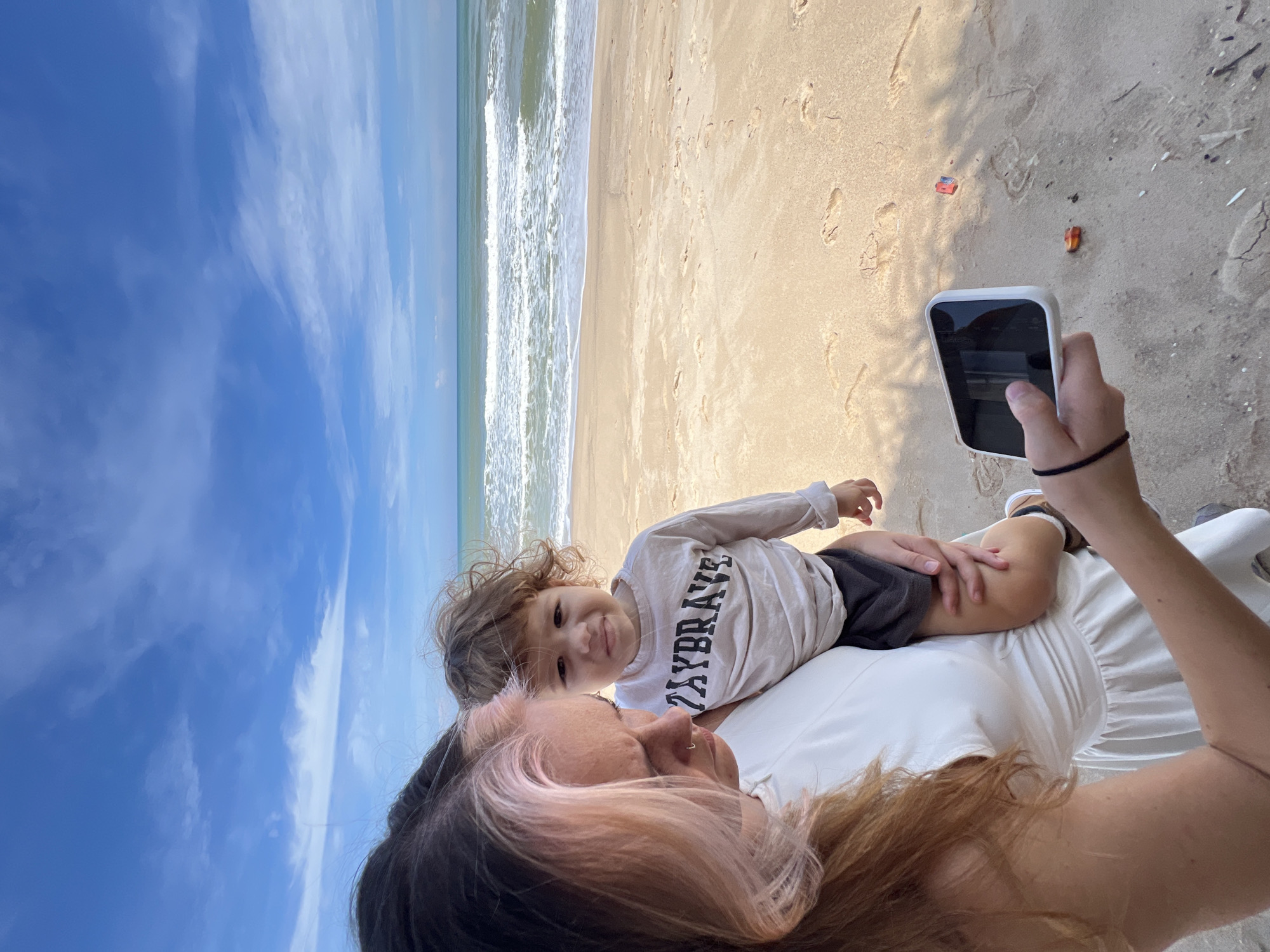 A woman with pink-streaked hair holds a young child on a sandy beach. The woman wears a white dress and is looking at a smartphone in her hand. The child, wearing a long-sleeved shirt with "PLAYBRAVE" printed on it and dark shorts, looks at the camera with a pouting expression. The ocean and sky are visible in the background. The scene is a beach, with footprints in the sand.