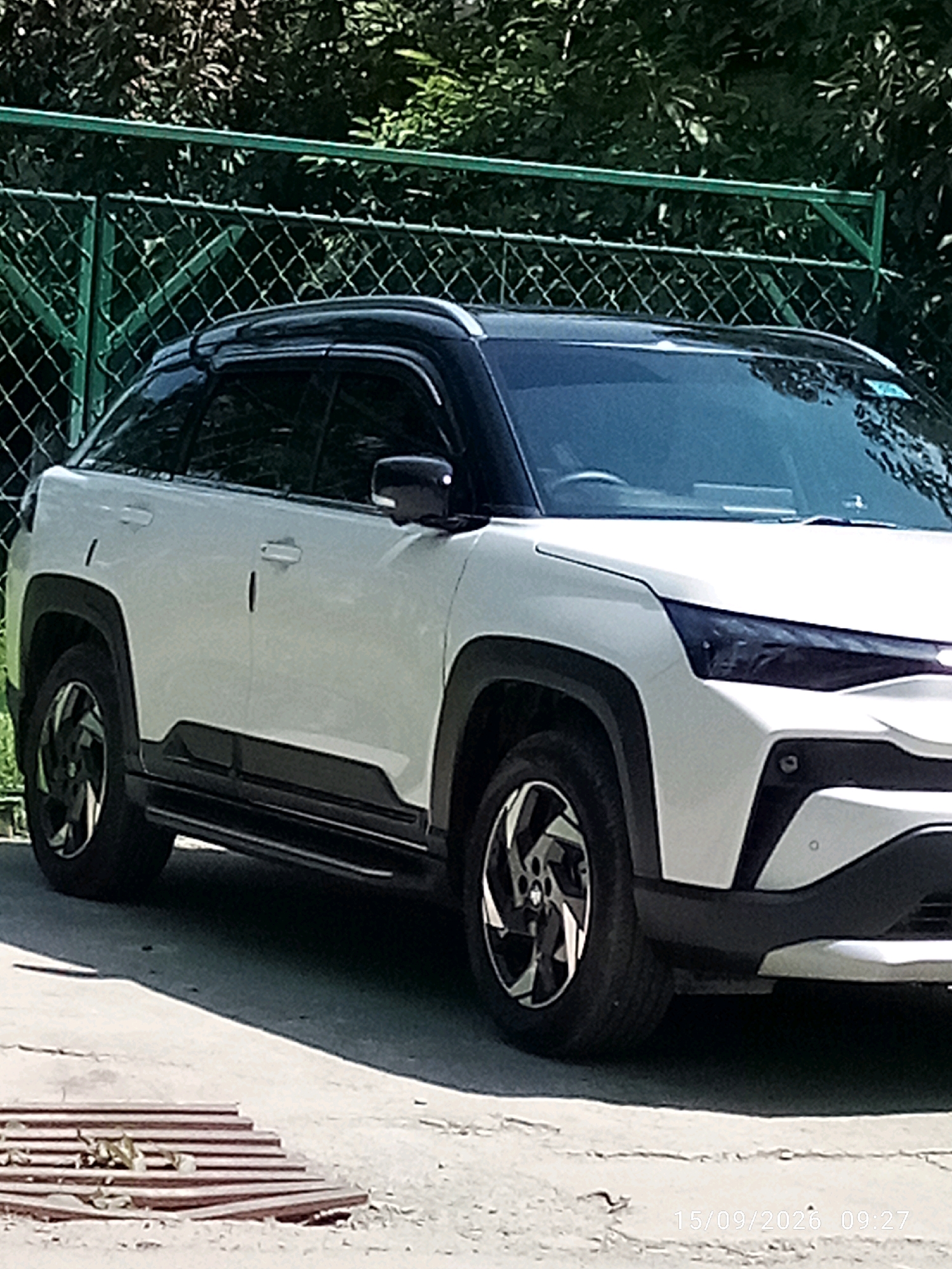 A white and black SUV is parked on a paved surface in front of a green metal fence and foliage. The vehicle has a two-tone paint scheme with a black roof and black trim around the wheel wells and lower body. The front of the vehicle shows a distinctive headlight design and a silver accent on the bumper. The wheels are black with a silver geometric spoke pattern. A wooden grate with some leaves on it is visible in the lower left corner of the image. The background consists of dense green leaves and trees. A date and time stamp, "15/09/2026 09:27", is visible in the lower right corner of the image. The scene appears to be an outdoor setting in Imphal, India.