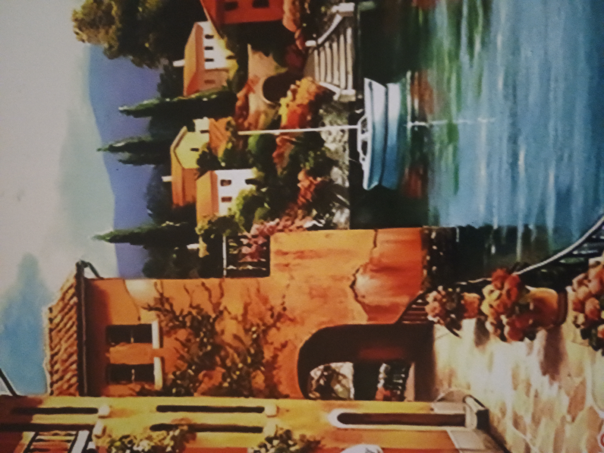 The image depicts a painting of a European town situated along a waterway. Buildings with terracotta facades are adorned with flowers and vines. In the background, a body of water reflects light, with a small sailboat visible on its surface. Other buildings are positioned further back, nestled among trees and greenery.