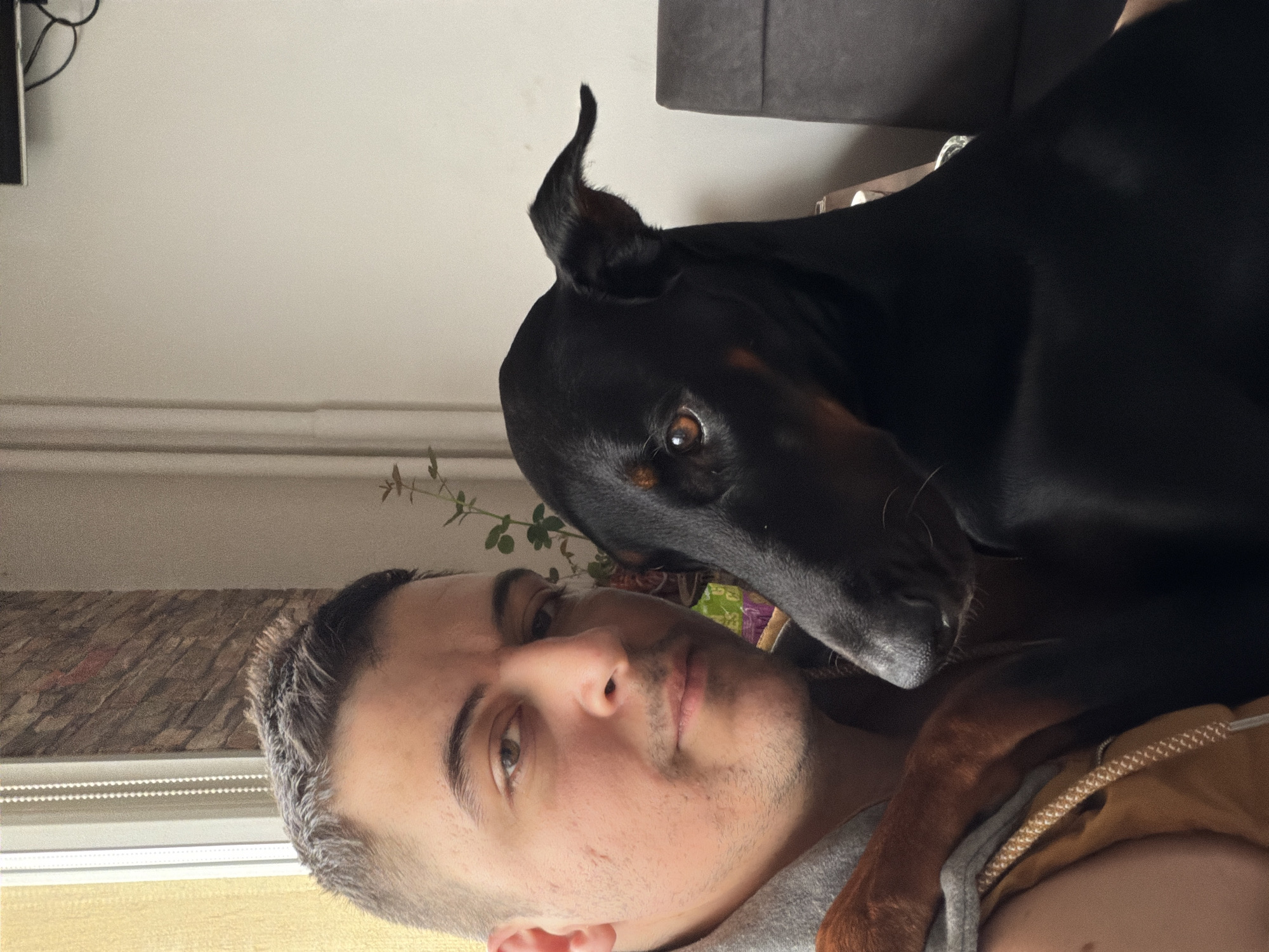 A man and a Doberman Pinscher dog are in a close pose, likely indoors. The man is light-skinned with short, dark hair, and is looking at the camera. He has a light beard. The Doberman is primarily black with brown markings on its muzzle and paws, and one ear is upright. The background includes a light-colored wall, a radiator, a partially visible window with blinds, and a section of a gray sofa. Due to lack of specific identifiers, the exact location in Sofia, Bulgaria cannot be determined.