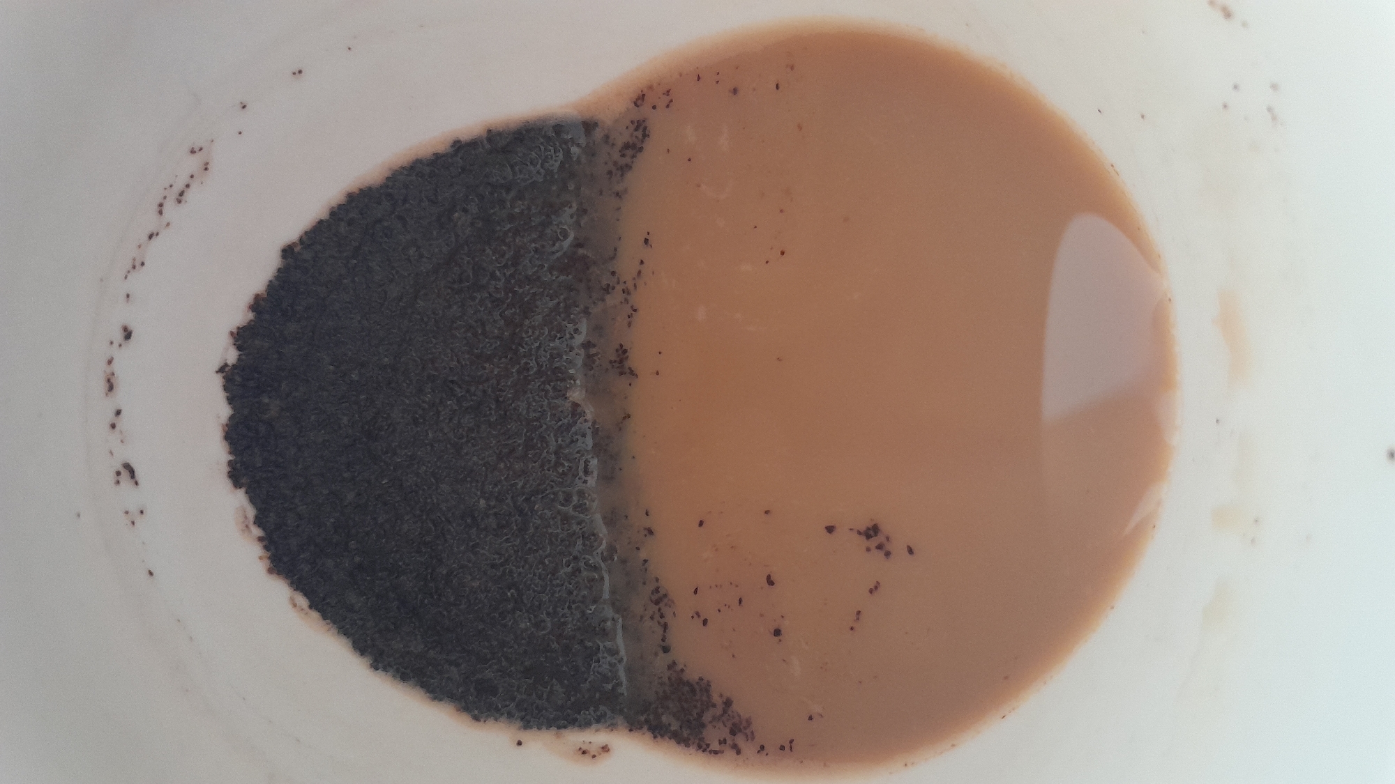 A close-up, overhead view of a white cup contains a beverage with loose tea leaves. The beverage is a light brown color, suggesting it is tea with milk. A thick layer of dark brown, granular tea leaves is visible on the top half of the cup, partially submerged in the liquid. Some tea leaves are also scattered in the liquid and along the rim of the cup. The lighting is bright, casting soft shadows. The image appears to be taken indoors, possibly in a kitchen. There are no people or other objects visible in the frame. The location is Unknown, Unknown.