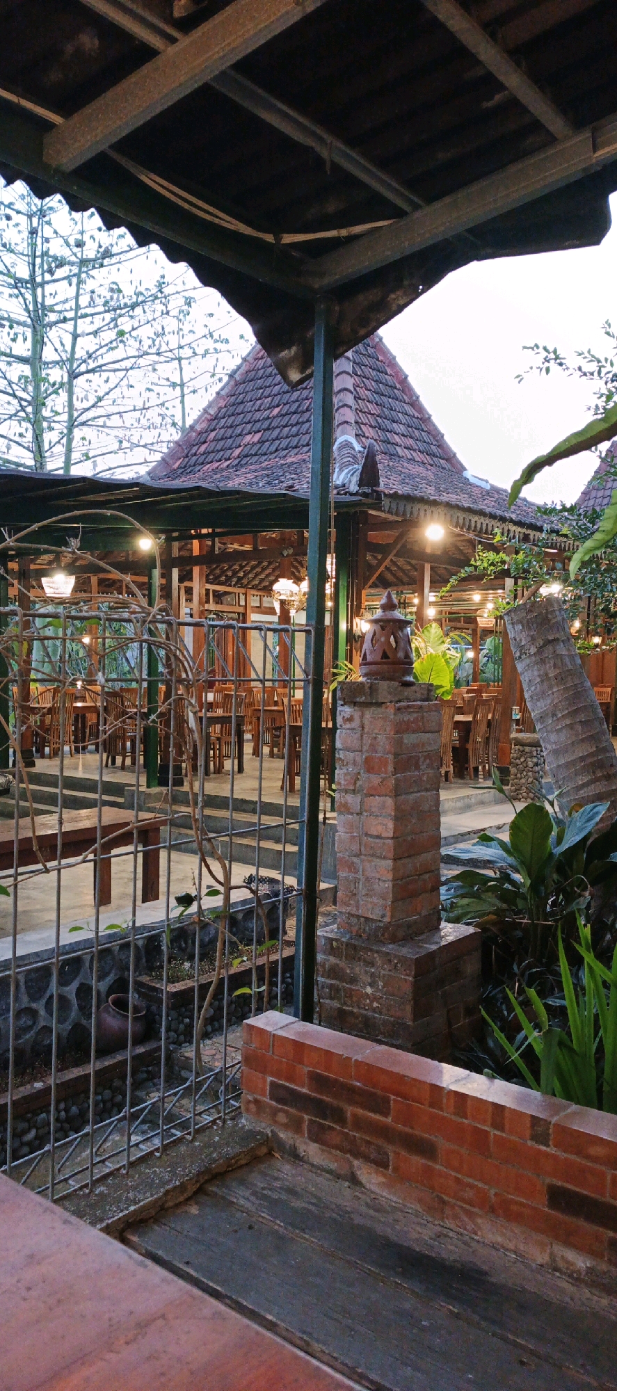 The image depicts an outdoor dining area of a restaurant, likely in Sidokarto, Indonesia, with a traditional architectural style. The setting features a pitched, terracotta-tiled roof supported by wooden beams and pillars. Numerous tables and chairs, made of dark wood, are arranged within the covered space. String lights and decorative lamps illuminate the area, suggesting it is evening or late afternoon.
In the foreground, a brick pillar supports a lantern with an intricate, cutout design. A metal fence, adorned with climbing vines, separates the foreground from the dining area. Beyond the fence, a low stone wall with a clay pot is visible. To the right of the brick pillar, a section of a palm tree trunk and lush green foliage are present. The ground is paved with concrete and some areas appear to be wooden decking. The overall atmosphere is relaxed and inviting, hinting at a cultural dining experience. No people are visible in the image.