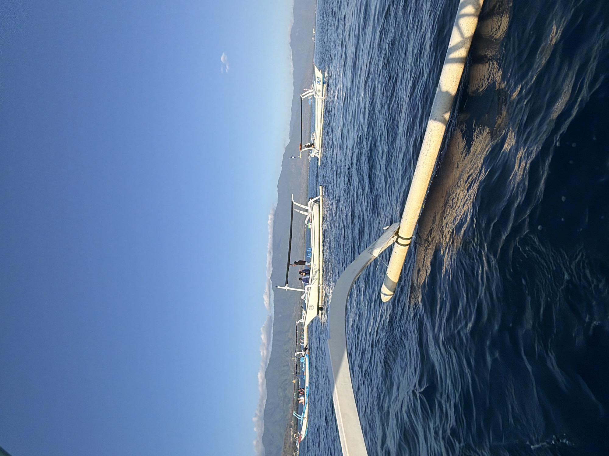 This image depicts a calm sea with several traditional Indonesian boats, possibly in Seririt, Indonesia. The sky is a clear, bright blue with a few wispy clouds. In the foreground, the edge of another boat is visible, with a white hull and a yellow outrigger pole. Further out on the water, three other boats are present, each with a thatched canopy and a few people on board. The boats appear to be moving slowly or anchored, and the water is a deep blue with gentle ripples. In the background, a range of mountains forms a hazy silhouette against the horizon, suggesting a coastal or island landscape. The overall scene conveys a sense of peacefulness and leisure on the water.