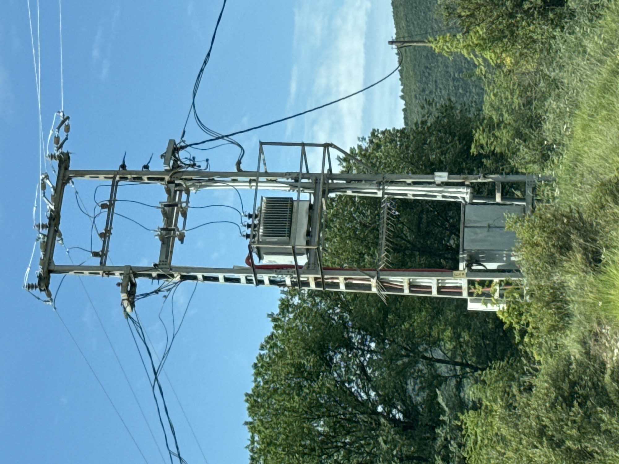 The image depicts a power pole structure located in a rural environment, seemingly in Podlastva (Poljice), Montenegro. The structure is made of metal and supports electrical transformers and insulators. Numerous wires extend from the pole, connecting to power lines. The surrounding environment is characterized by vegetation, including trees and bushes. A mountainous landscape is visible in the background under a blue sky with thin clouds.