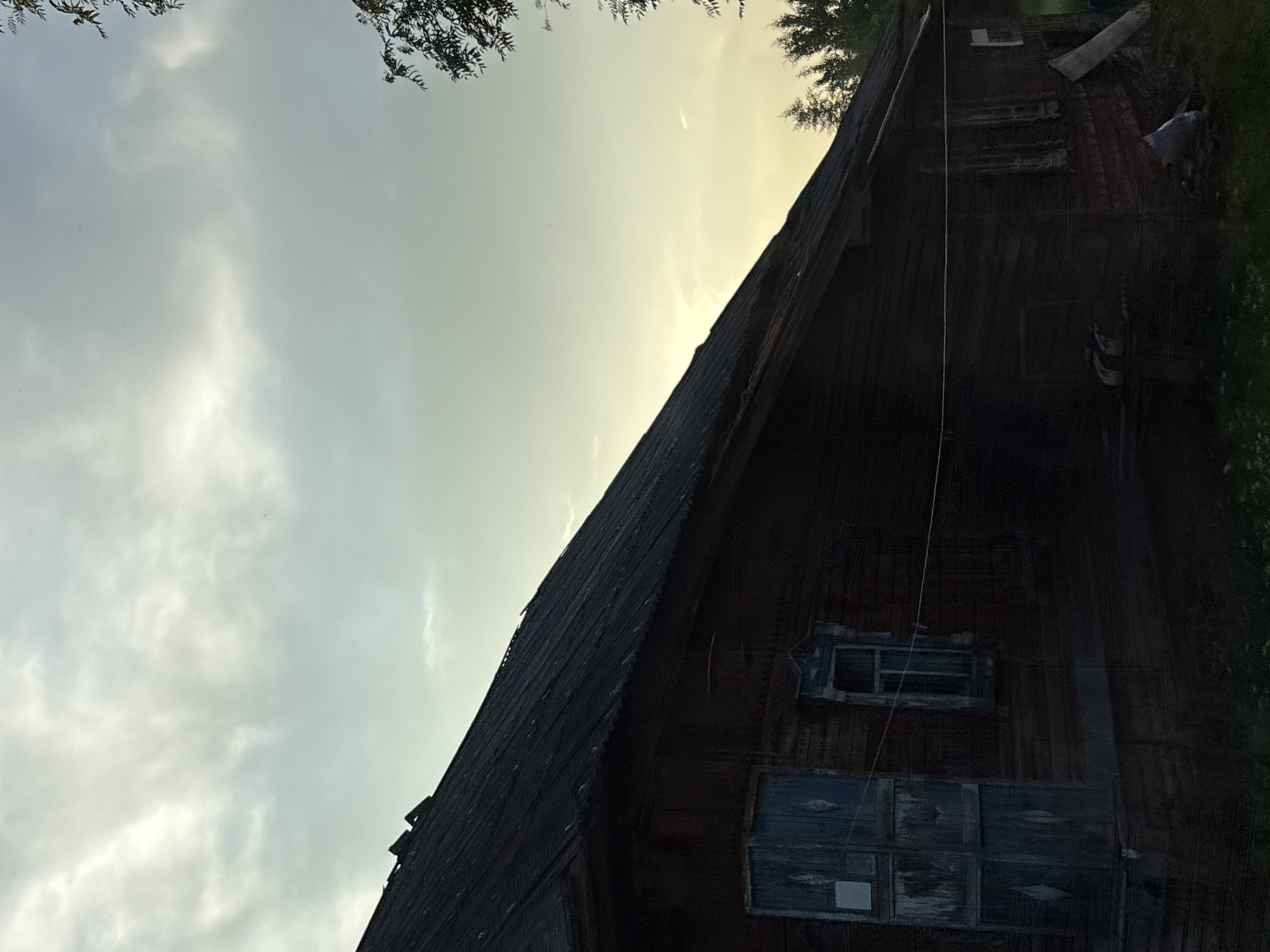 An exterior, eye-level, medium shot shows the side of a dark, weathered wooden house in Дресвяново, Russia, under a cloudy, pale blue sky. The house has a steeply pitched, dark roof and dark wooden siding. A pale blue, weathered door with visible grain and peeling paint is on the left side of the house. To the right of the door, a small, recessed window with white trim is visible. Further to the right, another small window with decorative white trim and a visible pane of glass is present. A thin white wire extends horizontally across the facade of the house. Trees and foliage are visible in the background on the right side of the frame. The ground in front of the house is covered with dark grass and some lighter green vegetation. In the lower right corner, there is a pile of debris including a white bag and a piece of wood. The overall impression is one of an old, rural dwelling.
