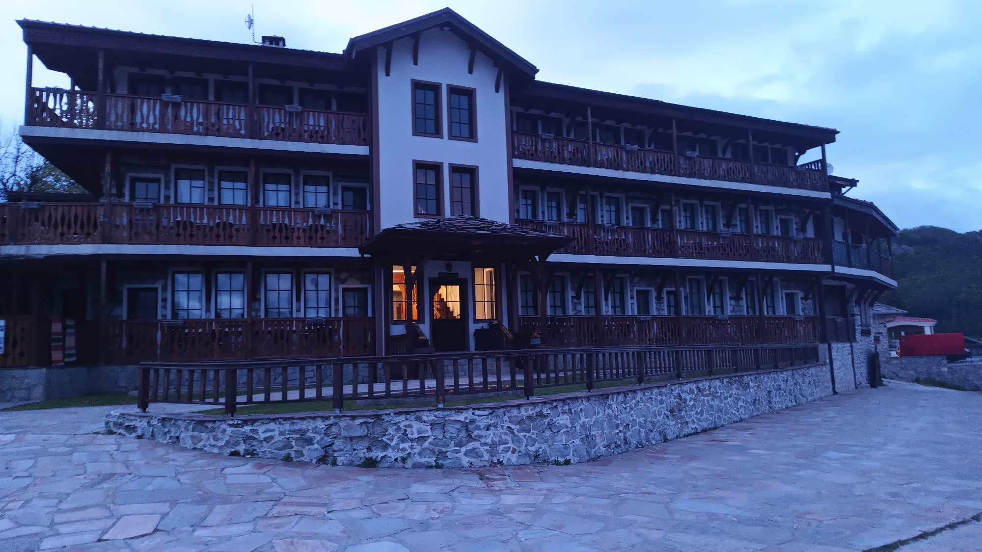 The image shows a multi-story building with a light-colored facade and dark brown wooden balconies. The building has multiple windows, and the entrance is illuminated, suggesting it is nighttime or dusk. A low stone wall with a wooden railing encloses a small garden area in front of the building. The ground in the foreground is paved with irregular stone slabs. The image is taken at the location of Vrata, Bulgaria. No people are visible in the image.