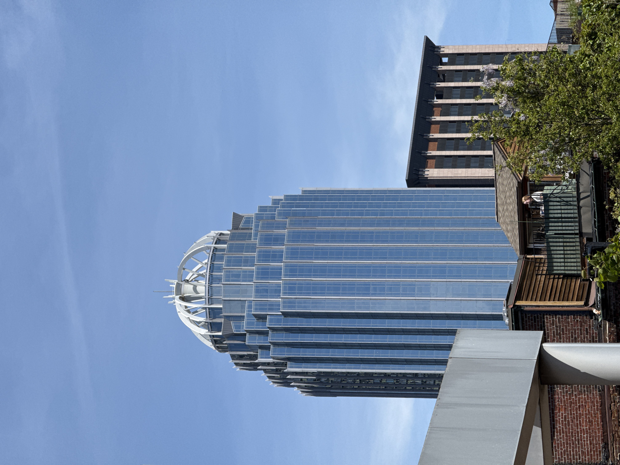 The image depicts a view of a tall, modern glass skyscraper in Boston, Massachusetts, United States of America, with a clear blue sky as the backdrop. The building is characterized by its stepped or tiered design, culminating in a rounded, skeletal white structure at the top. The reflective glass panels of the building appear blue due to the sky's reflection. To the right of the skyscraper, parts of other buildings are visible, including a taller, less reflective building with a darker facade. A section of a smaller building with brickwork and a balcony is also present, with greenery partially obscuring the view. The presence of a person on the balcony is noticeable. The overall perspective is looking upward at the buildings, which creates a sense of scale and emphasizes the height of the skyscraper.