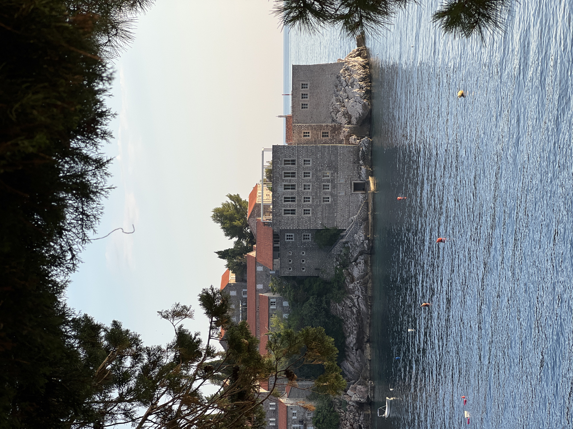 Framed by dark green pine branches, the image depicts a historic island complex in Sveti Stefan, Montenegro, bathed in the warm light of late afternoon or early morning. The complex, connected to the mainland by a narrow causeway, is characterized by its stone buildings with terracotta tiled roofs. The foreground is dominated by the calm, deep blue water of the Adriatic Sea, dotted with several buoys of various colors, including orange, yellow, and pink. A small white motorboat is moored near the rocky shore of the island. The island itself features a mix of fortified stone structures and older residential buildings nestled amongst lush green foliage. The clear sky above suggests fair weather, with a subtle haze on the horizon where the sea meets the sky. The overall scene evokes a sense of timeless beauty and tranquility.