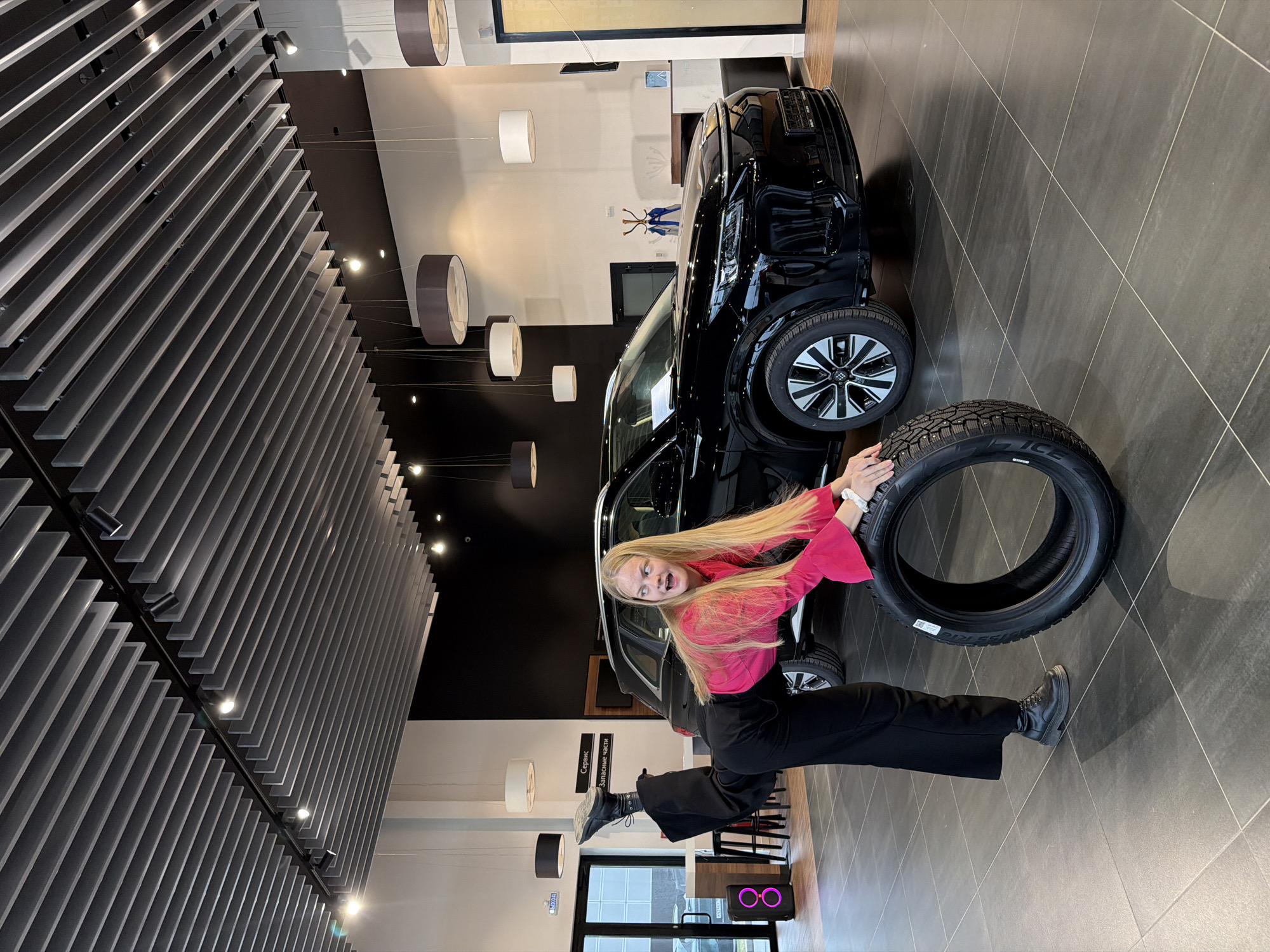 A young, fair-skinned woman with long blonde hair stands in a black shirt and black pants in a showroom in Izhevsk, Russia. She is standing next to a black sport utility vehicle and holding a black tire. A grey floor is beneath her. Overhead lighting is affixed to a black ceiling.