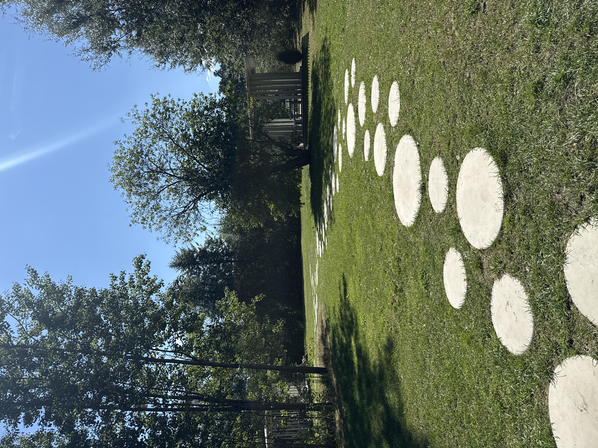 A daytime, eye-level, full-shot captures a circular stone pathway meandering through a manicured green lawn. The path starts in the foreground and curves towards a modern, open-air pavilion in the background, framed by tall trees on either side. The sky is a clear, bright blue with a sun flare visible in the upper right corner. The pavilion features a minimalist design with vertical white pillars supporting a flat roof and a table and chairs inside. The environment appears to be a park or a well-maintained garden, potentially located in Moscow, Russia, given the typical architectural style and landscaping for such areas in the city. No people are present in the scene.