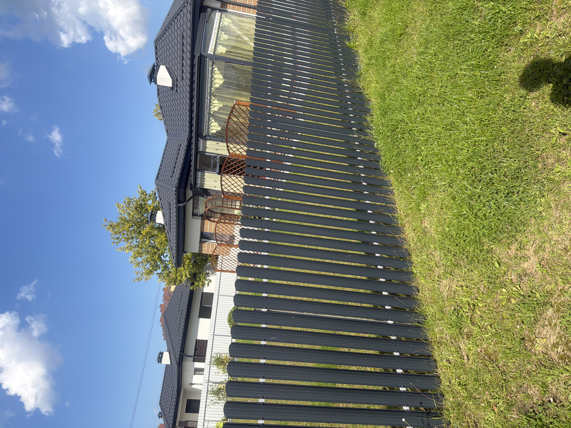 In Golubevo, Russia, a gray metal picket fence runs diagonally across the foreground, separating a grassy yard from a row of modern houses. The houses have white facades and dark gray tiled roofs, with chimneys visible on some. Behind the fence, the houses are partially obscured by greenery, including a tree with yellowing leaves. The sky is a bright blue with scattered white clouds. The scene depicts a residential area on a sunny day.