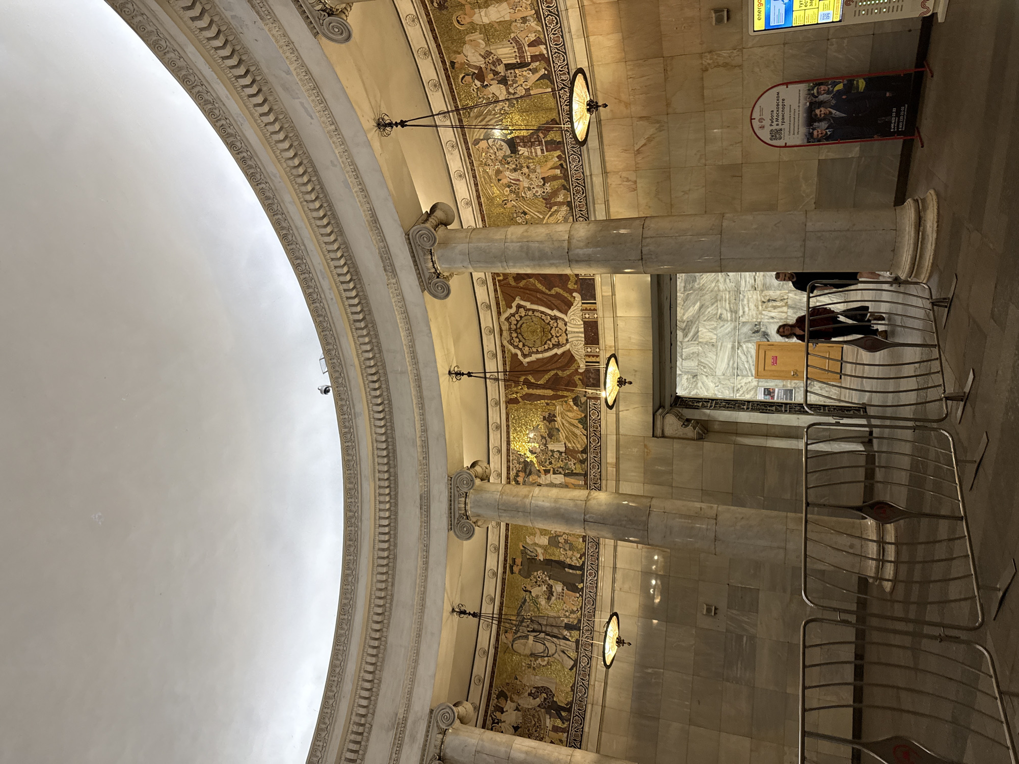 The image depicts the interior of a Moscow Metro station, specifically the entrance hall of the Komsomolskaya station. The architecture is grand, with a domed ceiling, large marble columns, and intricate mosaics adorning the upper walls. These mosaics feature scenes of people in traditional clothing, possibly depicting historical or cultural narratives. Four pendant lamps with ornate shades hang from the ceiling, illuminating the space. In the foreground, metal crowd control barriers are set up.
Two individuals, a man and a woman, are visible walking through an arched doorway towards the left of the frame. The woman wears a dark jacket and the man a black t-shirt. Further to the right, a recruitment poster for jobs in Moscow transport is displayed, featuring images of young men in professional attire and safety gear. Another digital display board, partially visible on the far right, shows yellow and blue signage. The walls are clad in large marble tiles, reflecting the ambient light. The overall impression is one of ornate and historical public architecture within a functional transit space.