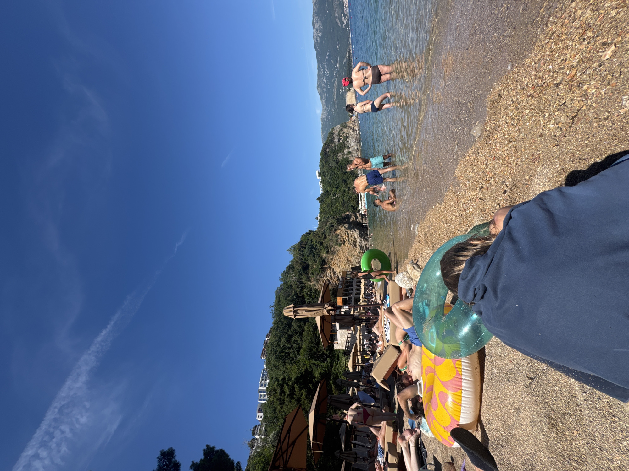 A bright, sunny day at a crowded beach in Budva, Montenegro. The scene is divided between the pebble beach and the clear, shallow water. On the beach, several people are sunbathing on lounge chairs under large umbrellas. In the foreground, two inflatable rings, one yellow and pink, the other green, lie on the pebbles. A person with their back to the camera is sitting near them, wrapped in a dark blue blanket or towel.
Further back on the beach, more people are scattered among lounge chairs and umbrellas. Behind the beach area, a picturesque building with an open-air structure, likely a restaurant or bar, is visible, nestled against a lush green hillside. Several more buildings are perched higher up on the slope, overlooking the scene.
In the water, a group of people are wading and interacting. Two men stand near each other, one in blue shorts and another in teal shorts. A woman in a black bikini stands facing them, while another woman in a brown bikini and red hat is further to the right. A person is also sitting in the shallow water to the far left.
The background features the azure blue sea meeting the horizon, with distant mountains under a clear blue sky. A thin, wispy cloud streak is visible in the upper portion of the sky. The overall impression is one of a lively summer day at a popular coastal resort.