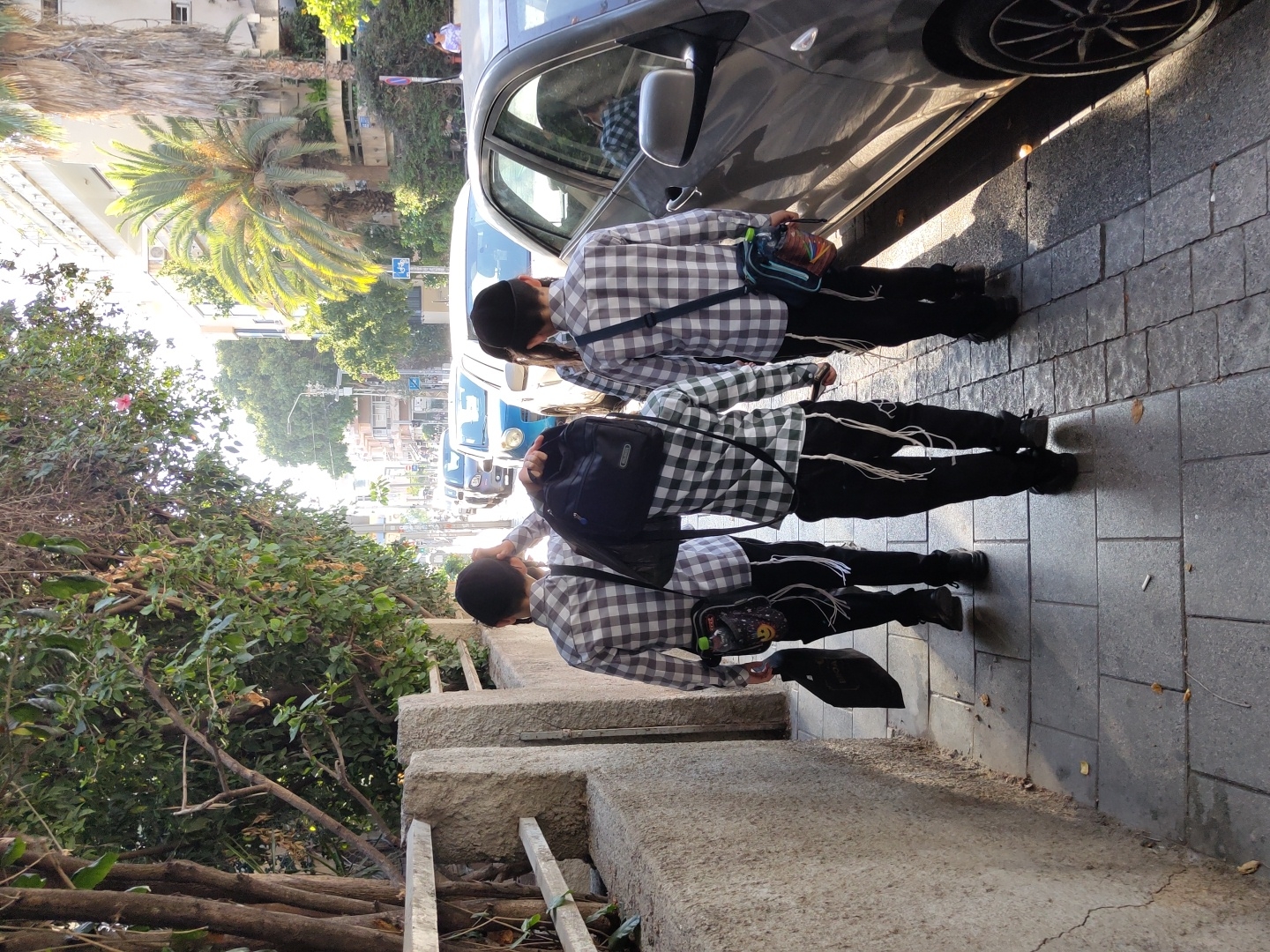 The image shows three young boys standing on a sidewalk in Tel Aviv, Israel. They are all dressed in checkered shirts and black pants, with yarmulkes on their heads. Two of the boys are wearing backpacks. The scene appears to be a sunny day, with palm trees and buildings visible in the background. The boys are walking alongside a parked car. There is no text visible in the image. The overall mood of the image is casual and ordinary, suggesting a normal day in the city. The lighting suggests it's daytime, likely morning or afternoon, given the shadows cast by the car.