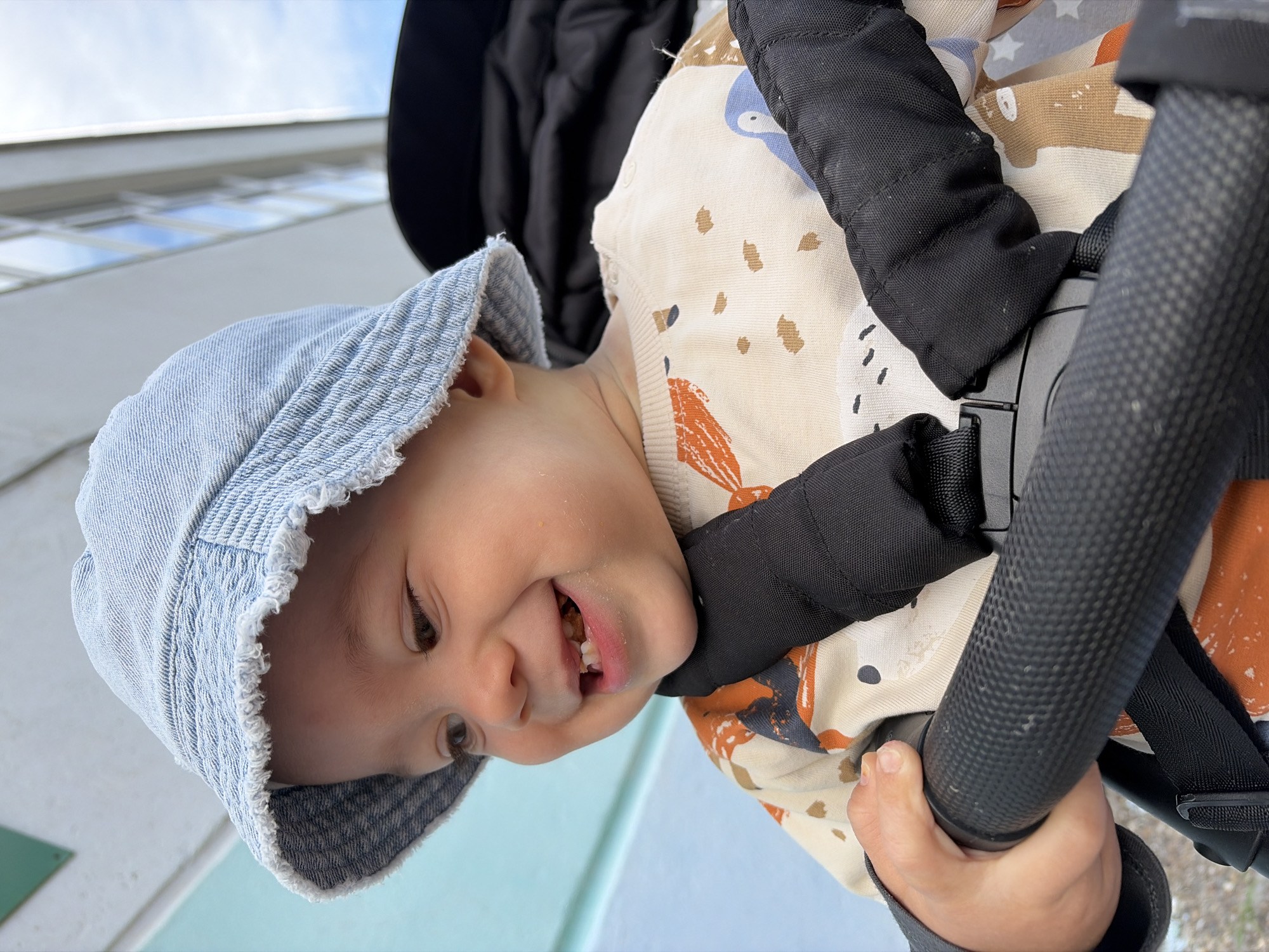 A young baby is pictured outdoors, seated in a stroller and looking towards the right side of the frame with a wide, joyful smile. The baby is wearing a light blue, denim-like bucket hat with a frayed brim and a cream-colored long-sleeved top adorned with a playful pattern of abstract shapes, dots, and what appear to be cartoon animals or figures in shades of brown, orange, and blue. Remnants of food are visible around the baby's mouth, and a few small teeth are showing.
The baby is secured in the stroller by a black harness system with padded straps and a central buckle. Their left hand is visible, gripping a black, textured bumper bar of the stroller.
In the background, a clear blue sky with some scattered white clouds is visible above a multi-story white building with a series of rectangular windows. A light blue-green surface is also present in the background, possibly part of the ground or another structure. The lighting indicates bright daylight. The scene captures a happy moment in Chaikovsky, Russia. No visible text is present.