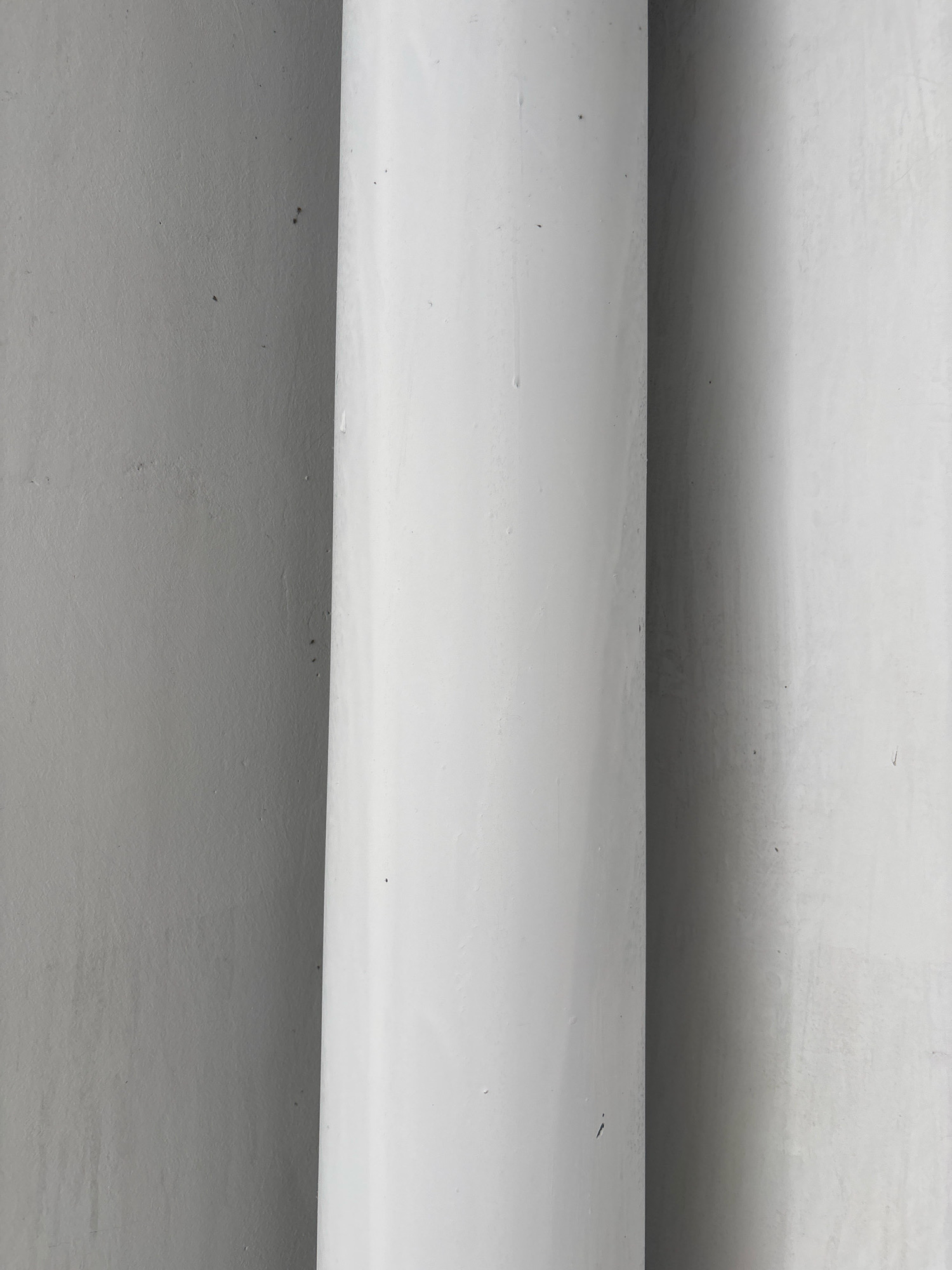 The image shows a close-up, abstract view of two large, white, cylindrical structures or pipes positioned vertically against a textured gray wall. The pipes appear to be made of a smooth material, possibly PVC or painted metal, and are casting shadows on the wall. The lighting suggests an indoor or shaded outdoor environment. The composition is stark and minimalist, focusing on the interplay of light, shadow, and texture. The image does not contain any identifiable landmarks, people, or specific objects that would indicate its location within Nha Trang, Vietnam, or any other city. The scene is devoid of any activity or interaction.