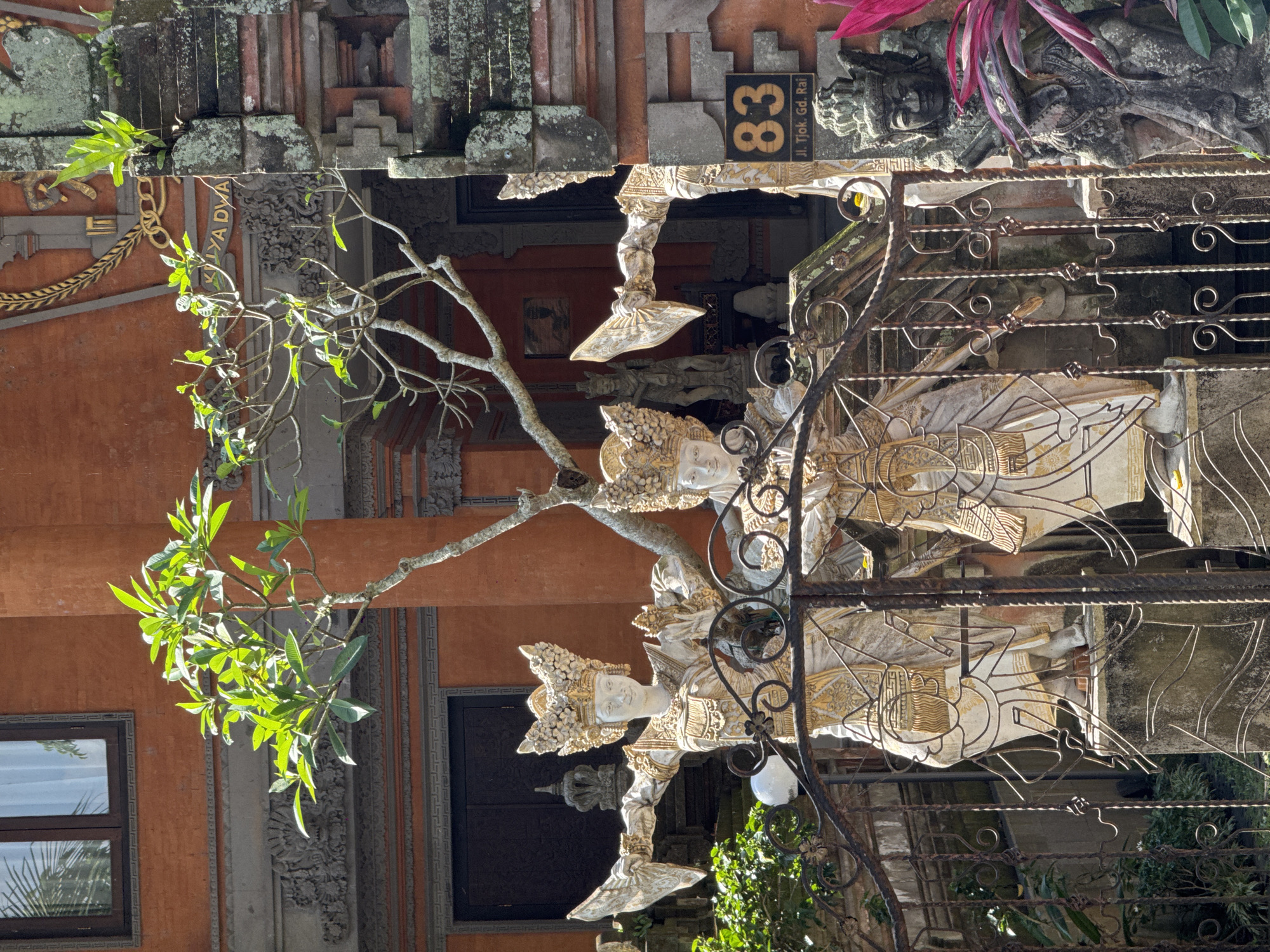 This image shows two ornate statues of Balinese dancers in front of a building with orange walls and intricate stonework. The statues are dressed in traditional attire with elaborate headdresses and gold accents. A tree with green leaves grows in front of them, with its branches reaching towards the building. A wrought iron gate separates the statues from the viewer. To the right of the statues, a stone statue of a guardian figure is visible, as well as a house number "83" with an address. The architecture and statues suggest a location in Bali, likely a temple or a traditional Balinese residence. The scene is bathed in sunlight, creating strong shadows. The overall impression is one of cultural richness and traditional artistry.