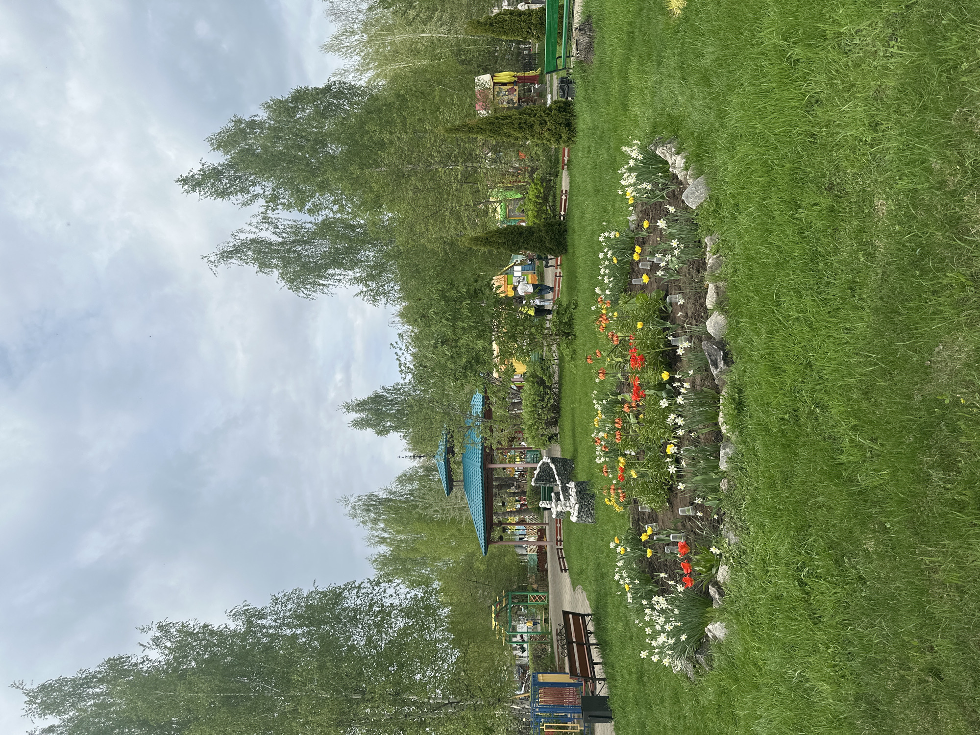 The image captures a vibrant park scene in an unidentified location. The foreground features a well-maintained lawn with a flower bed along its edge, showcasing a variety of colorful tulips and daffodils. A stone border separates the flowers from the grass.
Beyond the lawn and flower bed, there is a paved path that leads into the park, flanked by trees with light green foliage. Several benches are placed along the path. The path leads to a sheltered area with a unique blue tiled roof, supported by a structure.
Further into the park, there appear to be playground structures and art installations. The sky is overcast, with a soft light filtering through the clouds. No people are prominently visible in the image.