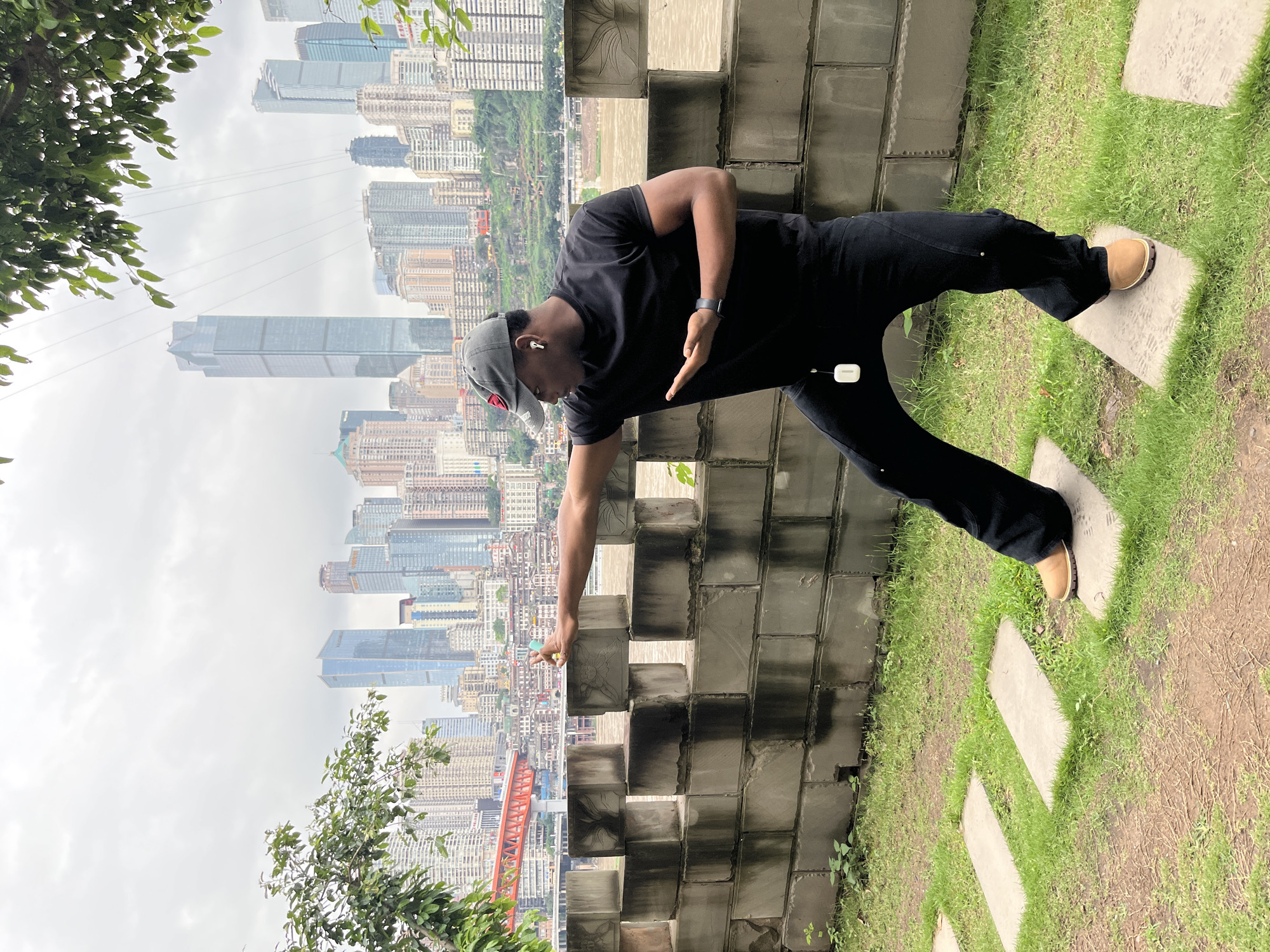 A man is standing on stepping stones next to a brick wall. He is wearing a black t-shirt and black pants and brown shoes. He is wearing a grey hat. The Yuzhong District skyline of Chongqing, China, is visible behind the wall. The sky is overcast.
