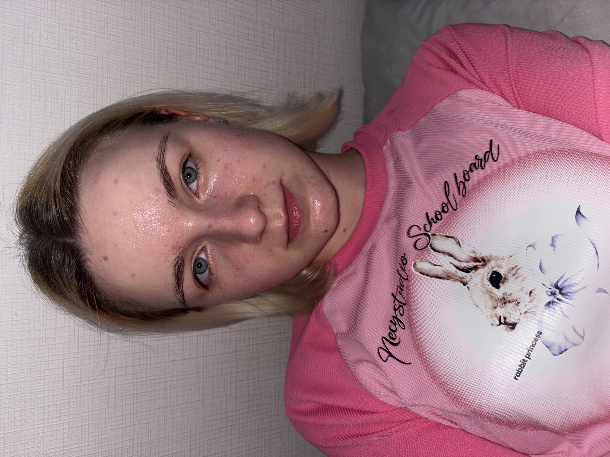 A slightly rotated close-up portrait shows a young individual with fair skin, light blonde hair with darker roots, and prominent blue eyes. The person looks directly at the camera with a neutral expression. Their skin appears somewhat shiny, possibly from moisture or oil, and exhibits several small red blemishes, particularly on the forehead and cheeks. A tiny, light blue stud earring is visible in their left earlobe.
The individual is wearing a two-toned pink long-sleeved top. The sleeves and upper portion of the shirt are a ribbed, solid pink. The central panel of the shirt is a lighter pink and features a circular graphic of a rabbit's head with a blue bow. Text is printed on the shirt: "Necystudio School board" is written in a black cursive font, and "rabbit princess" is written in a smaller, standard font below the rabbit graphic.
The background primarily consists of a light-colored, textured wall, likely wallpaper. To the right, a soft, light-colored object, such as a pillow or bedding, is visible, suggesting the person is lying down indoors. The lighting is even and soft, consistent with an indoor setting. The image captures a personal moment, likely a selfie, taken in a casual, residential environment.
