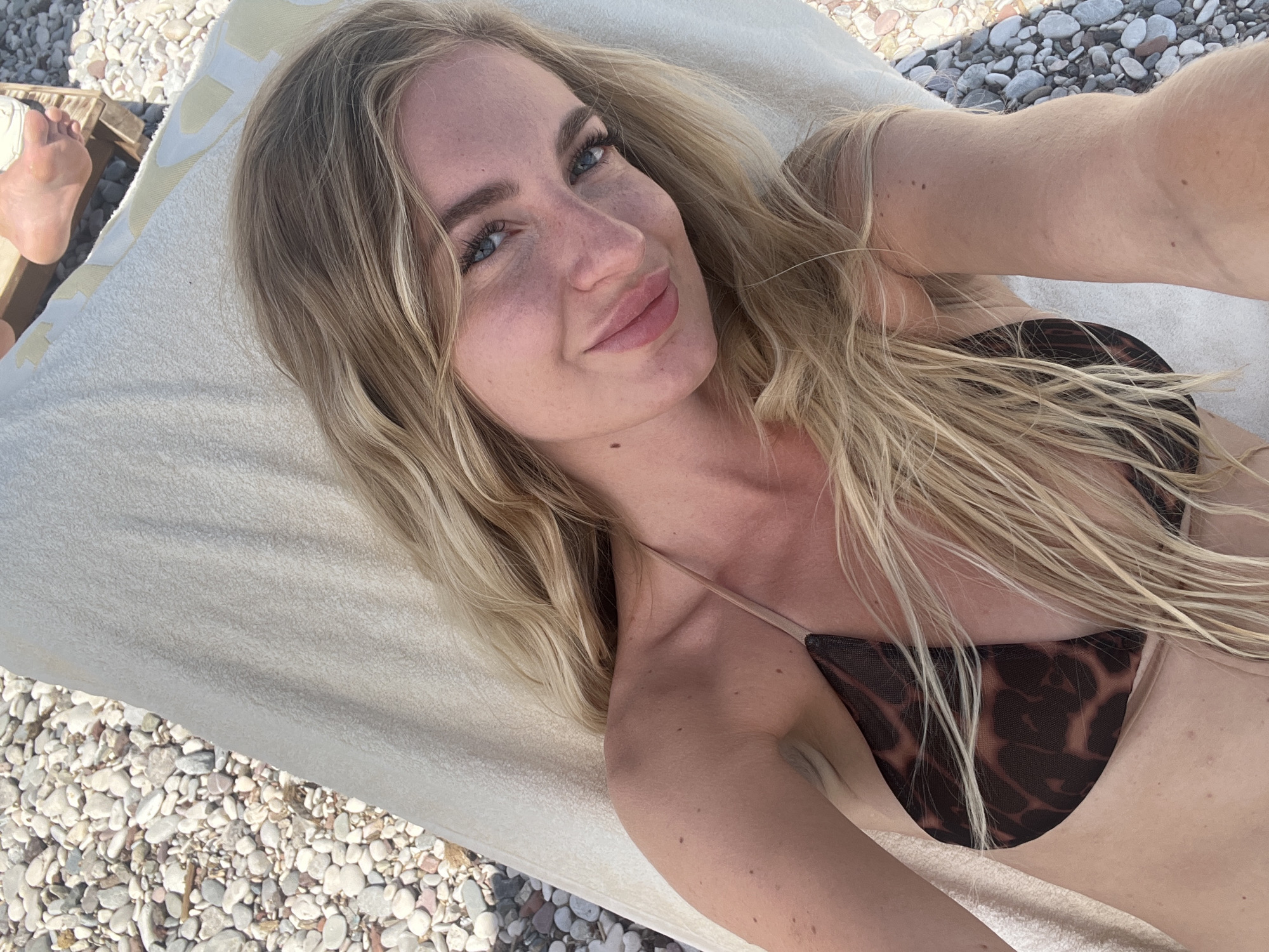 A fair-skinned woman is lying on a white towel in an outdoor setting. She has blonde hair, blue eyes, and is wearing a dark brown leopard print bikini top. Her left foot is visible in the upper left corner of the image, resting on a wooden pallet or chair. The ground appears to be covered in light-colored pebbles. Based on the setting, it is highly probable that the location is near a beach in Budva, Montenegro.