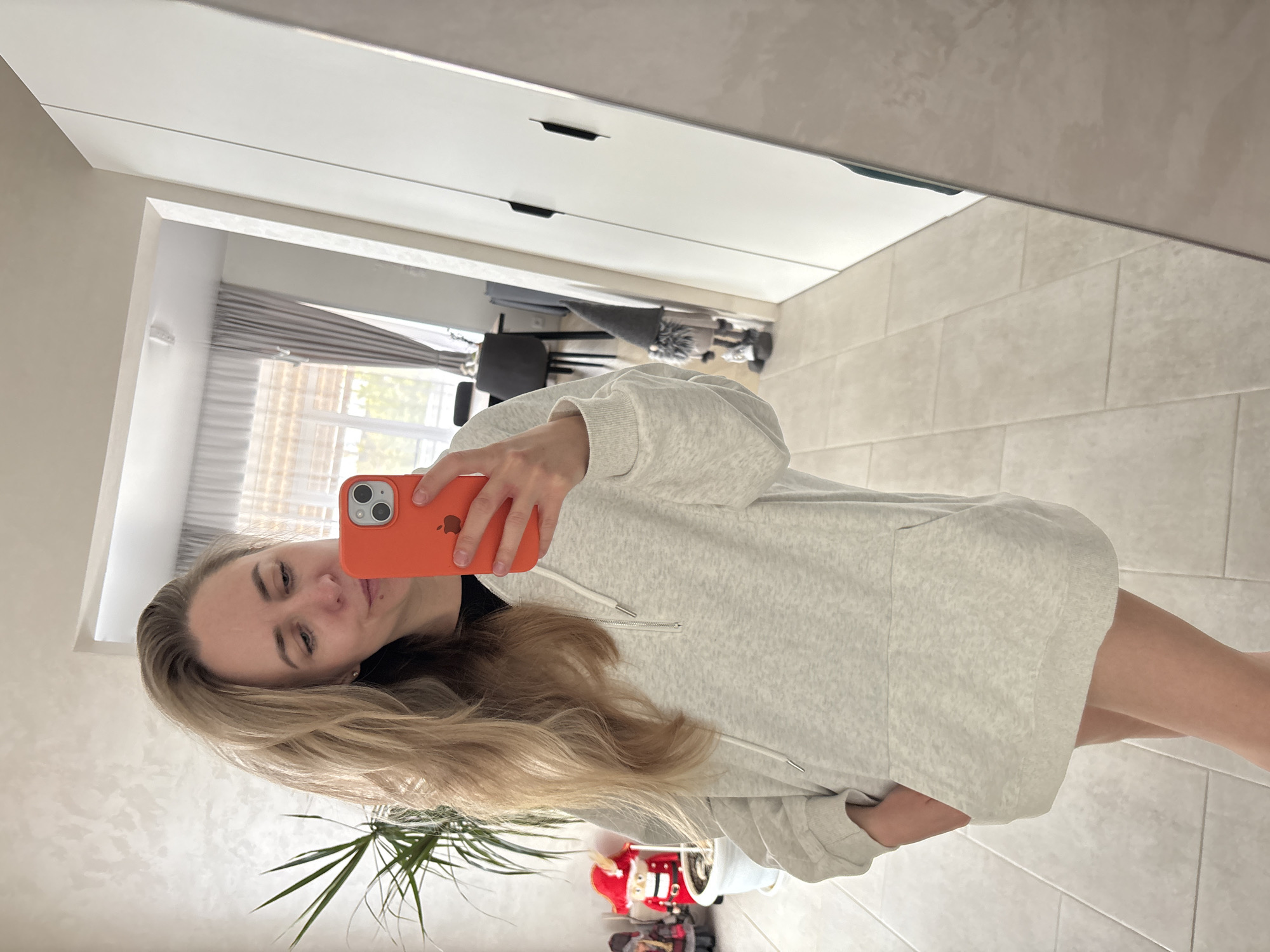 A woman is taking a selfie in front of a mirror. She is holding an orange iPhone. She is wearing a light gray hooded sweatshirt. Her blonde hair is long. The mirror reflects a room with tiled floors, a plant in the foreground, and furniture visible in the background. There is a small Christmas or holiday decoration at the base of the plant. The room appears to be in a residential setting. The location is Chaikovsky, Russia.