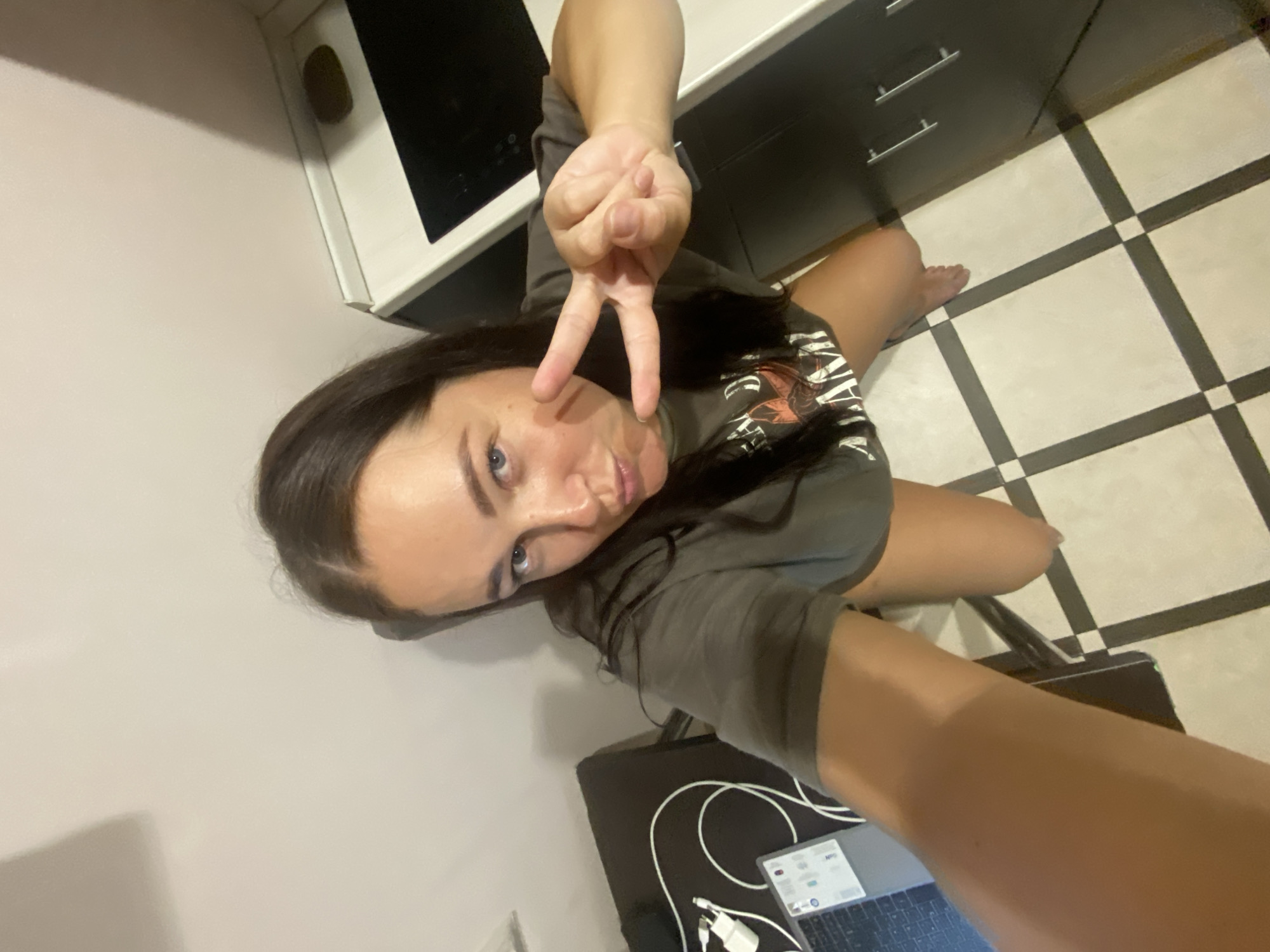 A woman with long, dark hair is kneeling on a checkered floor. She is wearing a dark t-shirt. Her right hand is raised, making a peace sign. She is looking at the camera with a playful expression. To her left is a laptop and some charging cables. In the background, there is a kitchen counter with a black stovetop. The scene appears to be an indoor residential setting. The image does not provide enough information to determine if it is located in Sochi, Russia.