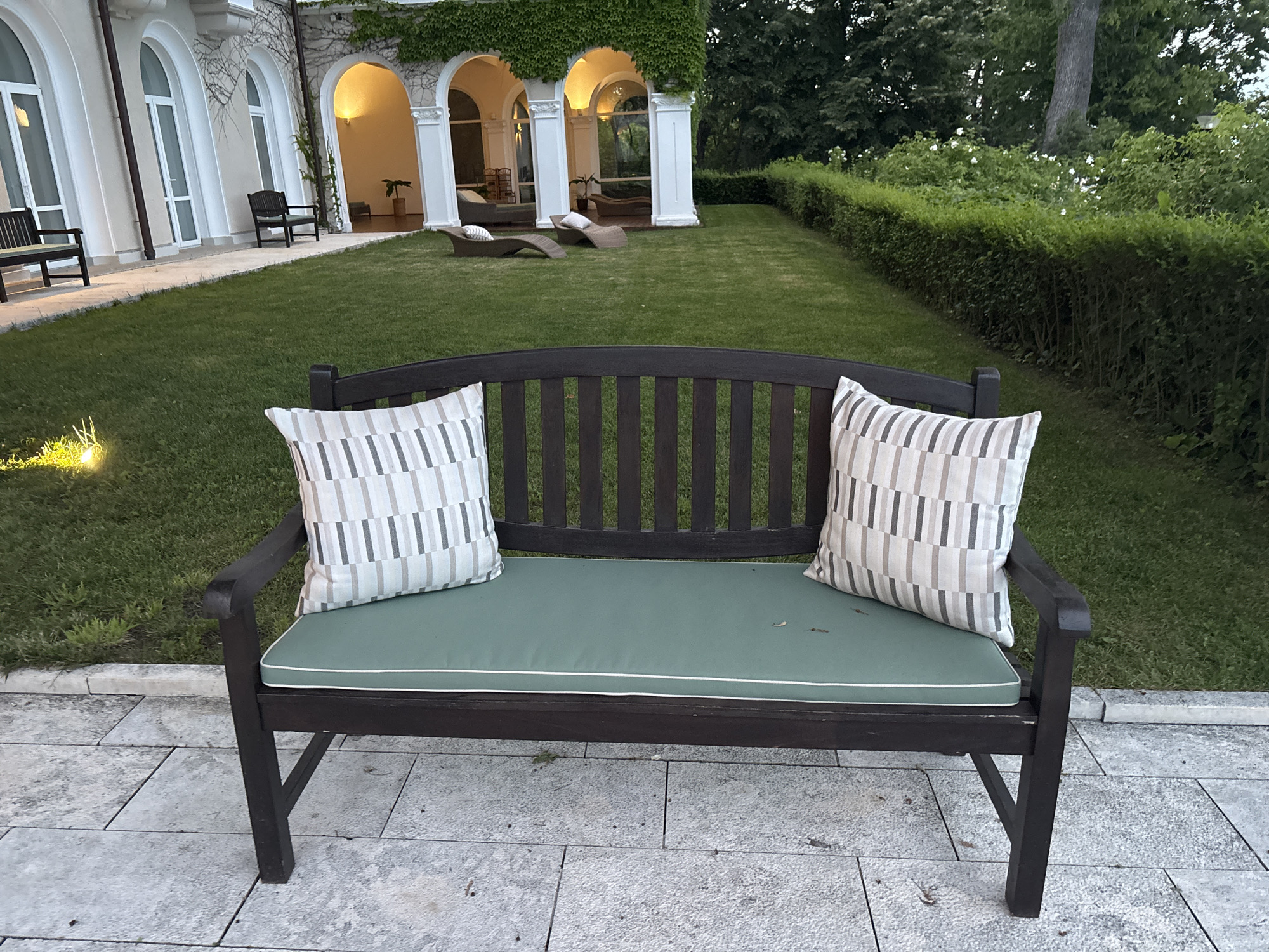 The image depicts an outdoor seating area in front of a building with arched windows. In the foreground, a dark wooden bench with a green cushion and two striped pillows sits on a stone patio. Behind the bench, a lush green lawn leads to a building with white arched doorways, some of which are illuminated by interior lights. One doorway reveals a glimpse of an interior space with potted plants and outdoor furniture. Arched windows with white frames are visible along the building's facade. A manicured hedge runs along the right side of the lawn, and mature trees form a backdrop. The scene suggests a tranquil setting, possibly a garden or courtyard associated with a residential or hospitality establishment. The image was taken in Varna, Bulgaria.