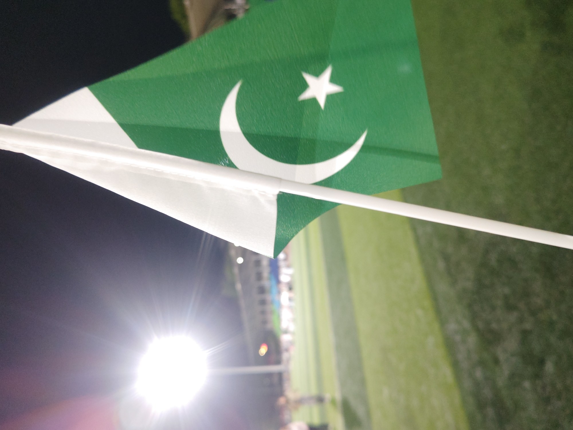 The image captures a Pakistan flag, green with a vertical white stripe on the hoist side, and a white crescent moon and a five-pointed star in the green field. The flag is held aloft by a white pole or stick, angled diagonally across the frame. A powerful, circular light source, likely a stadium floodlight, dominates the lower-left corner, casting a bright flare and illuminating the scene.
The background is predominantly dark, indicating nighttime, with a blurred expanse of green, suggesting a sports field or stadium turf. Faint, indistinct structures and blurry figures, possibly spectators or stadium personnel, can be observed in the distant background. The setting appears to be a nighttime sporting event or gathering in a stadium, where the Pakistan flag is being displayed, likely in a show of support or celebration. The event is situated in the Chongqing High-tech Zone, China. No visible text is present in the image.