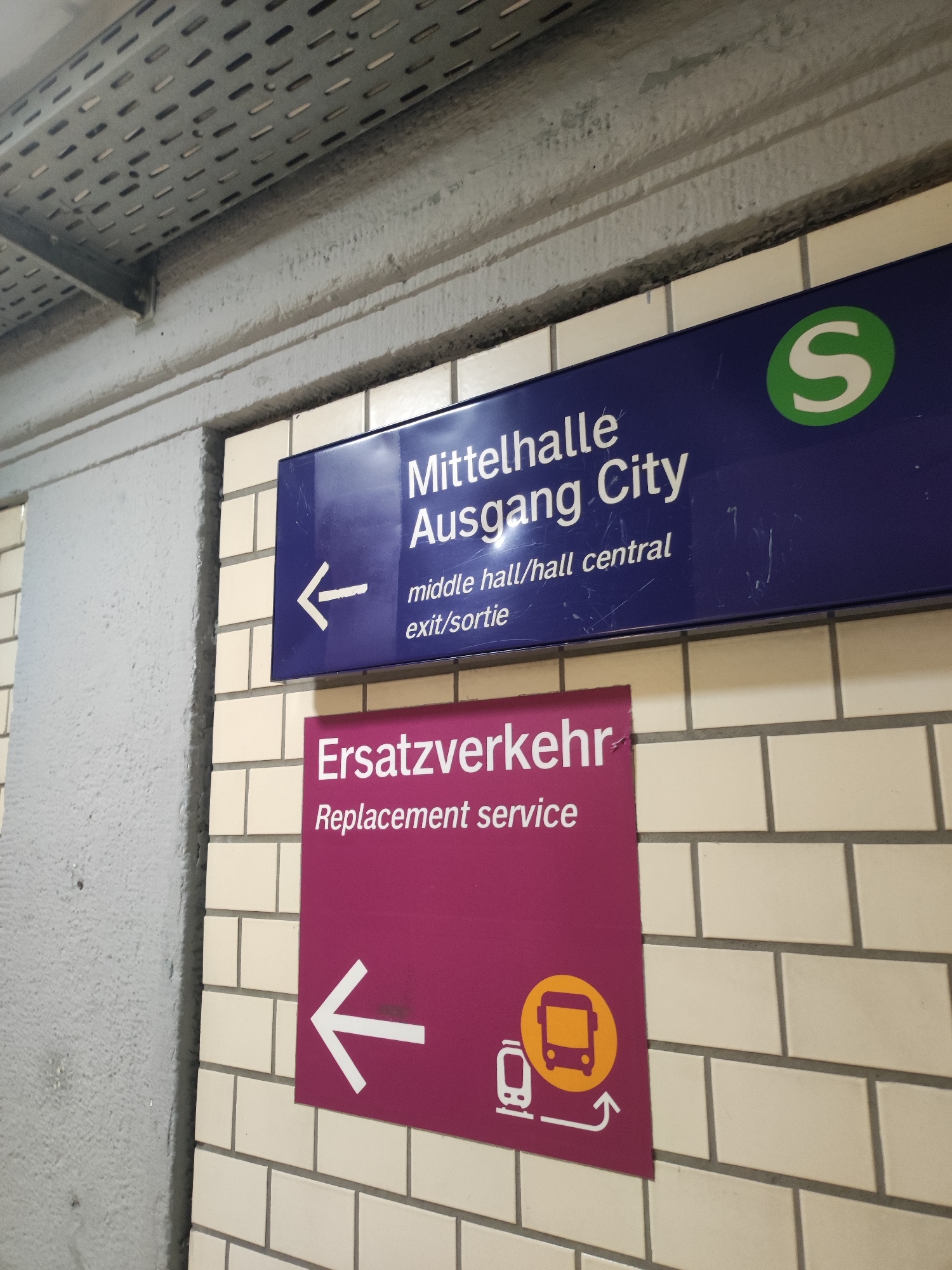 The image shows two signs mounted on a tiled wall. The upper sign is dark blue with white text and a green circular "S" logo. It reads "Mittelhalle Ausgang City" with smaller text below that says "middle hall/hall central exit/sortie". A white arrow pointing left is also visible on this sign. The lower sign is a deep pink with white text and icons. It reads "Ersatzverkehr Replacement service" and features a white arrow pointing left, accompanied by icons of a train and a bus within a yellow circle. The signs are located in an underground station, indicated by the ceiling infrastructure above and the tiled wall. This signage suggests a public transportation hub, likely a subway or S-Bahn station, in Nuremberg, Germany, as the "S" logo is associated with the S-Bahn system in Germany, and "Mittelhalle" and "Ausgang City" are German terms for central hall and city exit.