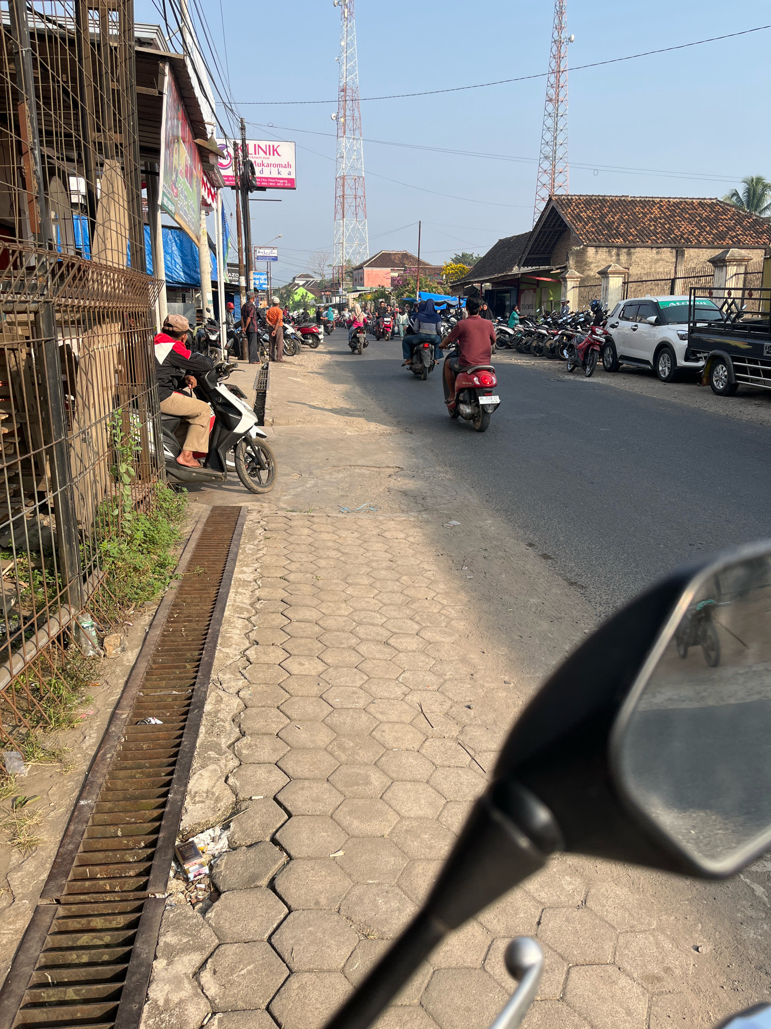 This image depicts a street scene in Kemuning, Indonesia. A man wearing a black and red jacket and tan pants is seated on a white motorcycle, parked alongside a sidewalk. To his right, a metal grate runs along the edge of the sidewalk. The street is busy with traffic, primarily motorcycles, with several people riding them. On the left side of the street, there are buildings with signs, including one for "Klinik Mukarromah" which appears to be a clinic. Across the street, buildings with tiled roofs are visible, and further down the road, two large cellular towers stand against a clear blue sky. In the foreground, a motorcycle's rearview mirror reflects the scene behind, showing more motorcycles and traffic. The overall environment suggests a typical day in a moderately busy urban or suburban area.